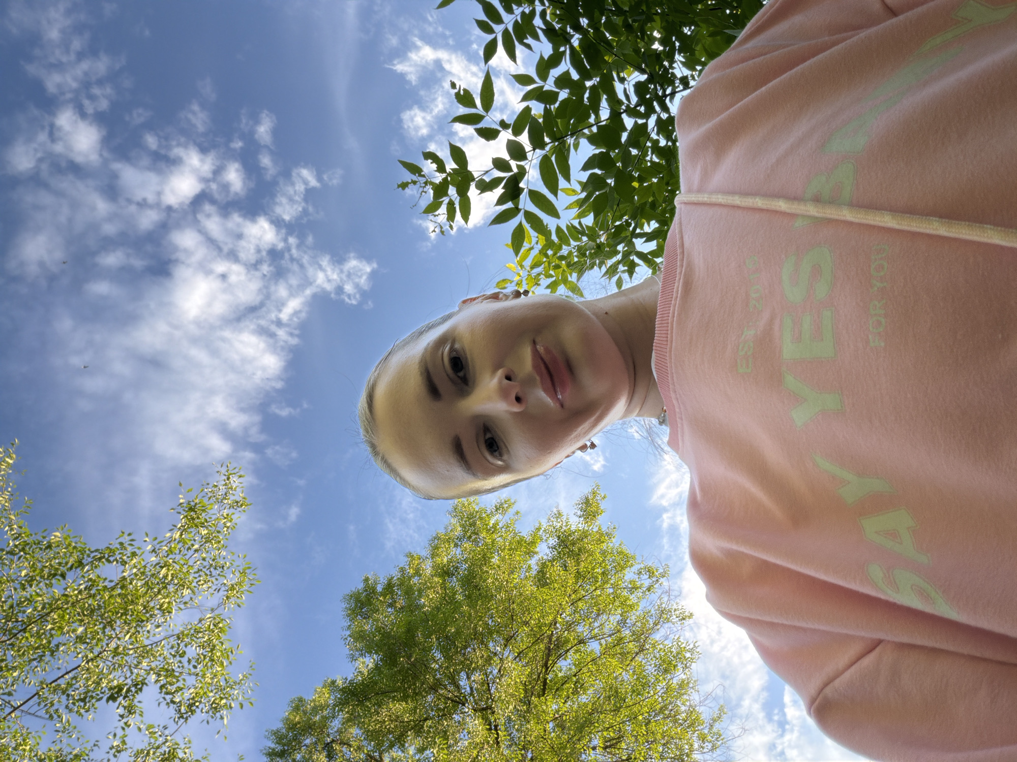 This is a low-angle, wide-angle photograph of a young woman looking up at the camera, taken outdoors during the daytime. The woman is positioned in the foreground, her face and upper chest visible. She has fair skin and blonde hair pulled back, and she is wearing a light pink hooded sweatshirt. The sweatshirt has a pastel green graphic on it that reads "SAY YES" in large letters, with "EST. 2019" and "FOR YOU" in smaller print below.
The background is dominated by a bright blue sky with scattered white, fluffy clouds. Green foliage from trees frames the top and sides of the image, suggesting a park or natural setting. Sunlight filters through the leaves, creating a dappled effect. The perspective implies the photo was taken from a low position, possibly lying on the ground or held above the head, looking upwards. The overall mood is bright and cheerful, with the woman's gaze directed towards the viewer. The presence of trees and open sky points to a warm weather season. The location context provided is Semiluki, Russia.