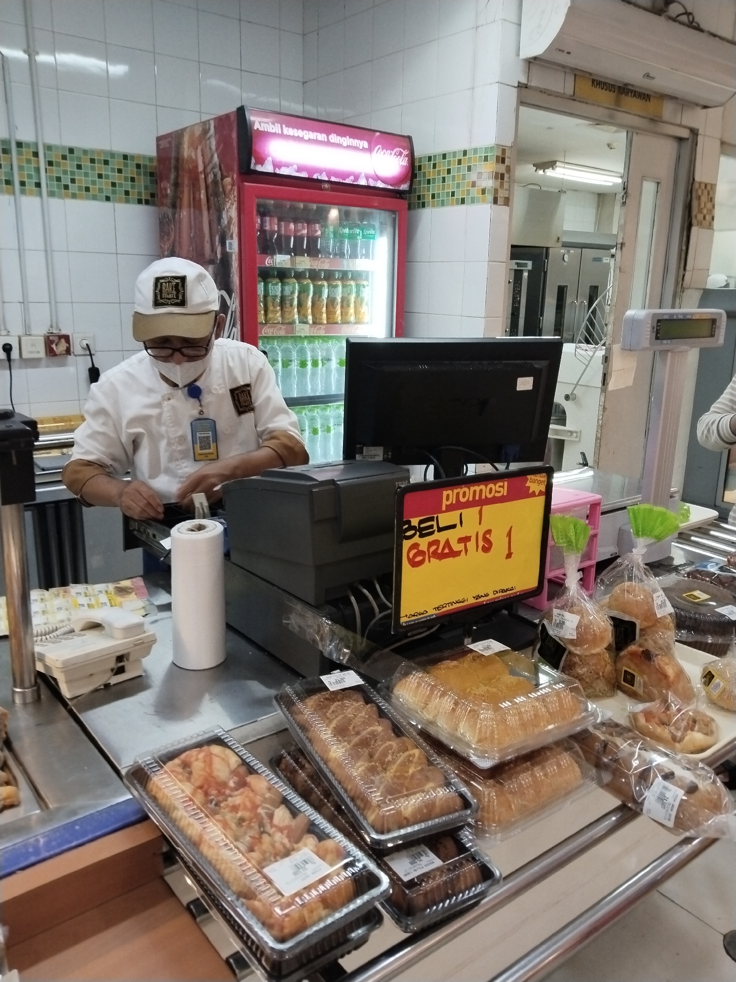The image shows an indoor scene, likely a bakery or a section of a supermarket in Kudus, Indonesia. A person wearing a white chef's coat, a white baseball cap, glasses, and a face mask is standing behind a counter, appearing to be attending to a transaction or preparing something. They are wearing a blue lanyard with an ID card.
The counter area is filled with baked goods in clear plastic containers and plastic bags. Prominently displayed on the counter is a yellow sign with red lettering that reads "promosi" (promotion), "BELI 1 GRATIS 1" (Buy 1 Get 1 Free), and "HARGA TERTINGGI YANG DIBAWA" (Highest price brought). To the left of the sign is a computer monitor and a cash register.
Behind the person, a red beverage refrigerator is visible, stocked with bottles of drinks. In the background, there is a doorway labeled "KHUSUS KARYAWAN" (Employees Only), suggesting a back area or staff-only space. The environment appears to be a commercial setting with tiled walls and shelving. The lighting suggests an indoor artificial light source.