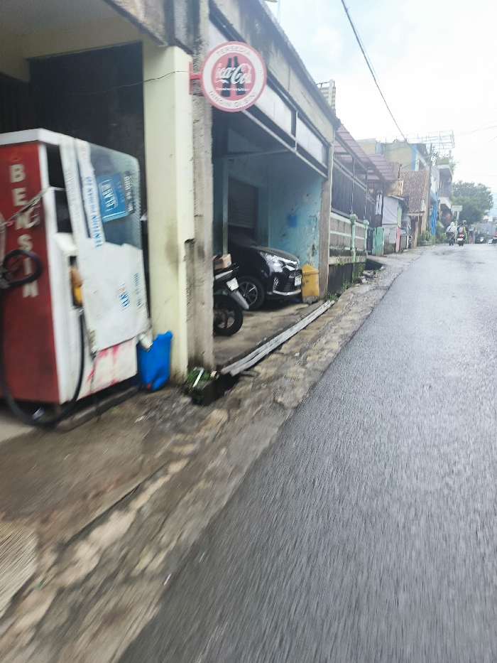 The image depicts a street scene in Cimenyan, Indonesia (latitude: -6.889, longitude: 107.651). A red and white gasoline pump sits on the edge of a paved street, partially obscured by a building. A black car is parked in front of the building, facing away from the viewer. A motorcycle is parked beside the car. A blue container sits near the pump. The street is wet and appears to have recently rained. Several buildings line the street on the right side of the image. A Coca-Cola sign hangs above the entrance to the building where the gasoline pump is located. The street is mostly empty, with no people visible. The buildings appear to be residential. The environment is overcast and cloudy.