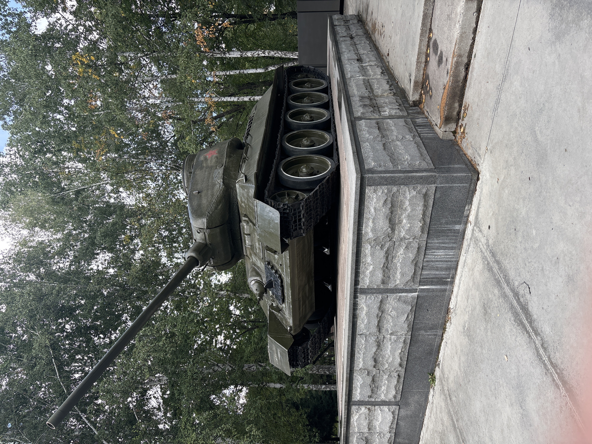The image shows a Soviet T-34 tank monument on display outdoors. The tank is painted olive green and is positioned on a concrete and stone pedestal. Behind the tank, a grove of trees with green and yellowing leaves is visible, suggesting an autumn season. The ground in front of the monument is paved with concrete. Based on the context and common knowledge of such monuments, this appears to be a war memorial. Specific details about the location in Novosibirsk, Russia, are not discernible from the image itself. There are no people or actions depicted.