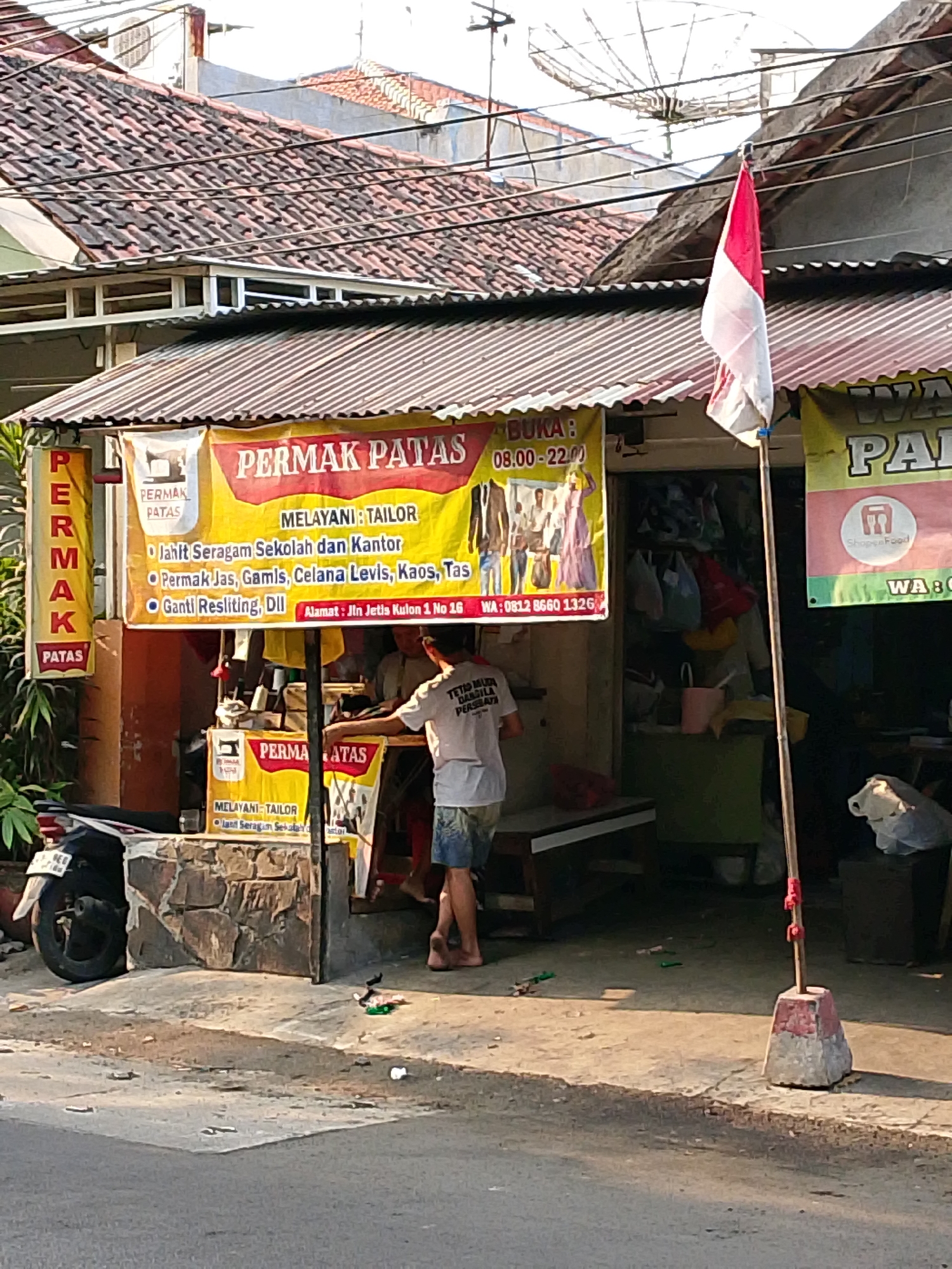An outdoor medium shot captures a tailor shop in a bustling urban environment, likely in Surabaya, Indonesia, given the context clues of a red and white flag, reminiscent of the Indonesian flag. The shop, identified by a prominent yellow banner advertising "PERMAK PATAS" and "MELAYANI: TAILOR," displays a variety of services including school uniforms, jeans, gamis, levis, kaos, and bags, with alterations and zipper repairs also offered. The shop operates from 8:00 AM to 10:00 PM.
A man, possibly the tailor, stands near the entrance, wearing a white t-shirt and shorts, his back partially to the camera, suggesting he is attending to a customer or an ongoing task. Another individual, seen from the chest down, is also visible inside the shop. A red and white Indonesian flag is prominently displayed on a wooden pole in front of the shop, catching the sunlight.
The scene is cluttered with items typical of a street-side business: a motorcycle is parked to the left of the shop, a wooden bench is positioned near the entrance, and various bags and clothing items are visible hanging inside and around the shop. The ground in front of the shop is paved, with scattered debris indicating a busy, lived-in space. Rooftops of residential buildings, covered in clay tiles, are visible in the background, with electrical wires crisscrossing overhead. A satellite dish is also perched on one of the rooftops. The overall impression is one of a functional, everyday business operating within a residential urban setting.