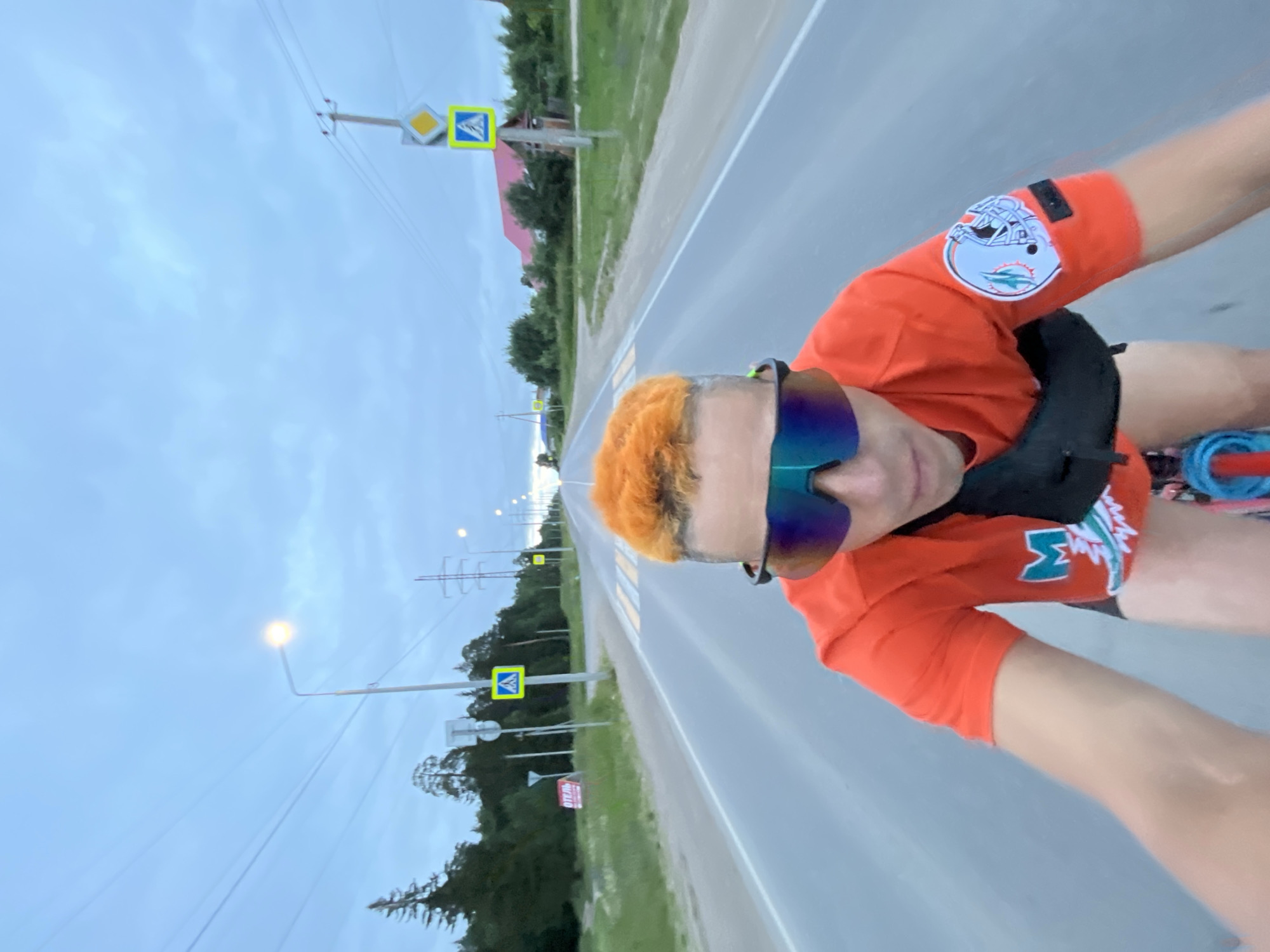 This media file shows a person with bright orange hair and blue-green sunglasses riding a bicycle down a paved road. The person is wearing an orange t-shirt with a white and blue logo on the left sleeve and a white "M" shaped logo on the right side of their shorts. The road appears to be in a rural or suburban area, with grassy verges, trees, and utility poles visible on either side. Several traffic signs are also present, including a pedestrian crossing sign. The sky is overcast, suggesting it might be evening or a cloudy day. The overall mood appears to be relaxed and active, with the rider seemingly enjoying their bike ride. The location is Chaikovsky, Russia. There is no explicit text visible in the image, aside from the logos on the clothing and traffic signs.