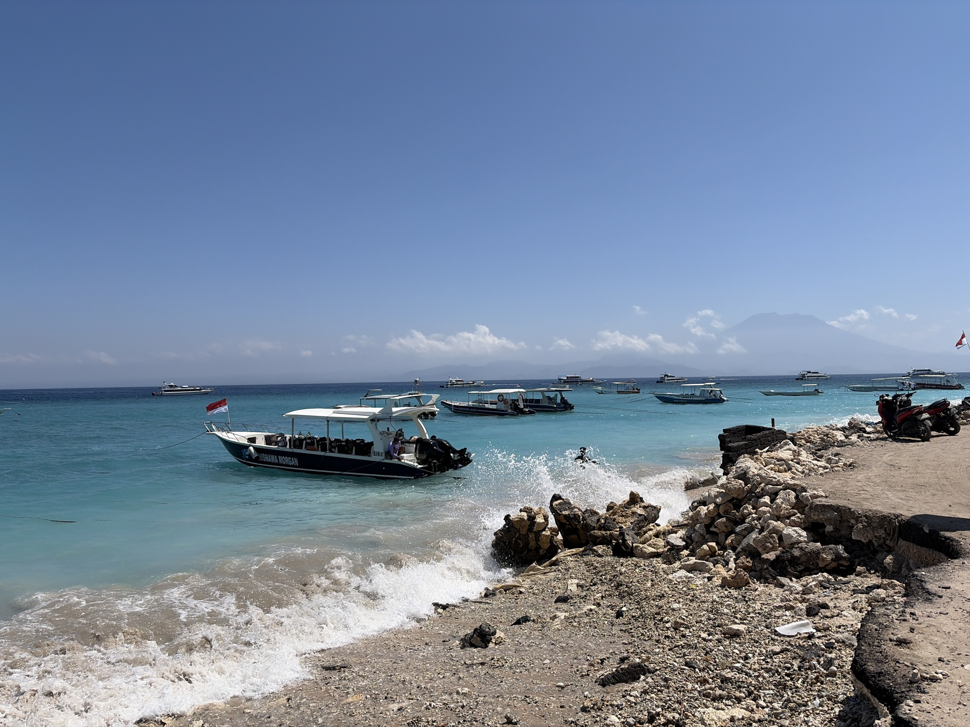 In Nusa Penida, Indonesia, a busy beach scene unfolds under a clear blue sky. A dark blue and white boat, branded with "NISNAWA MORGAN," is anchored in the turquoise waters, its passengers enjoying the day. The boat is equipped with an outboard motor and a canopy for shade. Several other boats are visible in the distance, scattered across the sea. The shoreline is a mix of sand and pebbles, with a rough, rocky embankment bordering a paved path. Waves are crashing onto the shore, creating a foamy spray. A red motorbike is parked on the paved area, suggesting local transportation. The overall impression is a vibrant tropical setting with activity on and near the water.
