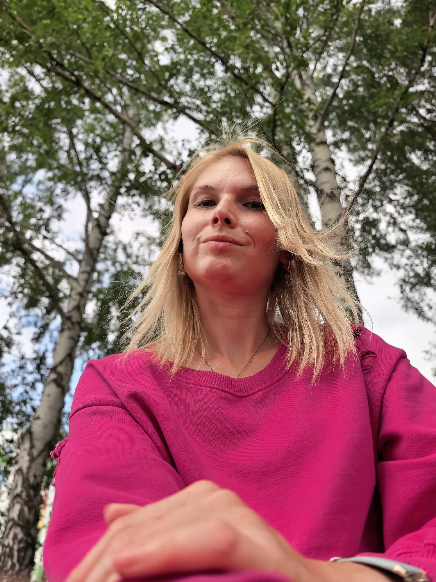 A low-angle, medium shot captures a young Caucasian woman with blonde hair and a smirking expression, wearing a bright pink sweatshirt, positioned outdoors amongst birch trees. Her hair is windswept, and she has a delicate gold necklace visible. The background consists of the white trunks of birch trees with green foliage and a cloudy sky. The woman's hands are visible in the foreground, one resting on her lap and the other slightly blurred in the lower right corner, adorned with a wristwatch. The image appears to be a selfie. The specific location in Sterlitamak, Russia cannot be determined from the visual information provided.