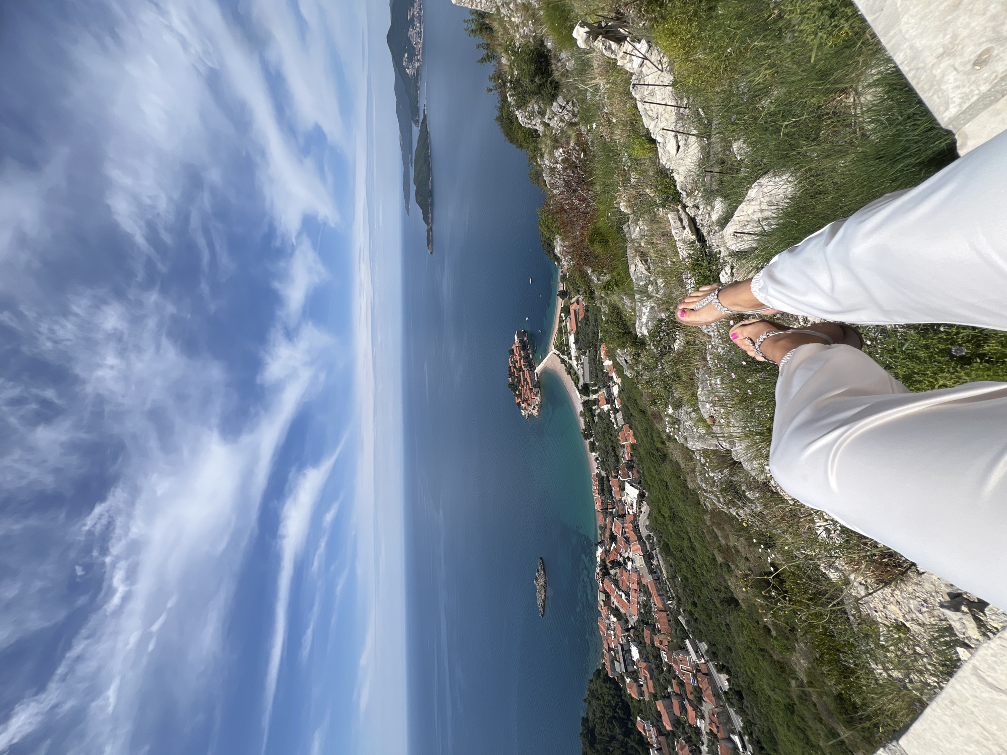 The image shows a high-angle view in Sveti Stefan, Montenegro. The shot is taken from a high point, likely a cliff or elevated platform. A person wearing white pants and sandals with pink painted toenails is partially visible on the right side of the image. They appear to be sitting or standing on the edge of the cliff.
Below the cliff, there's a view of the Adriatic Sea with scattered islands. The island of Sveti Stefan is visible; it is densely built with reddish-roofed buildings. The coastline below the cliff shows a beach with buildings extending inland. The sky is blue with light, wispy clouds. The cliff edge is covered with sparse grass and some vegetation. A section of a metal safety rail runs along the cliff edge.