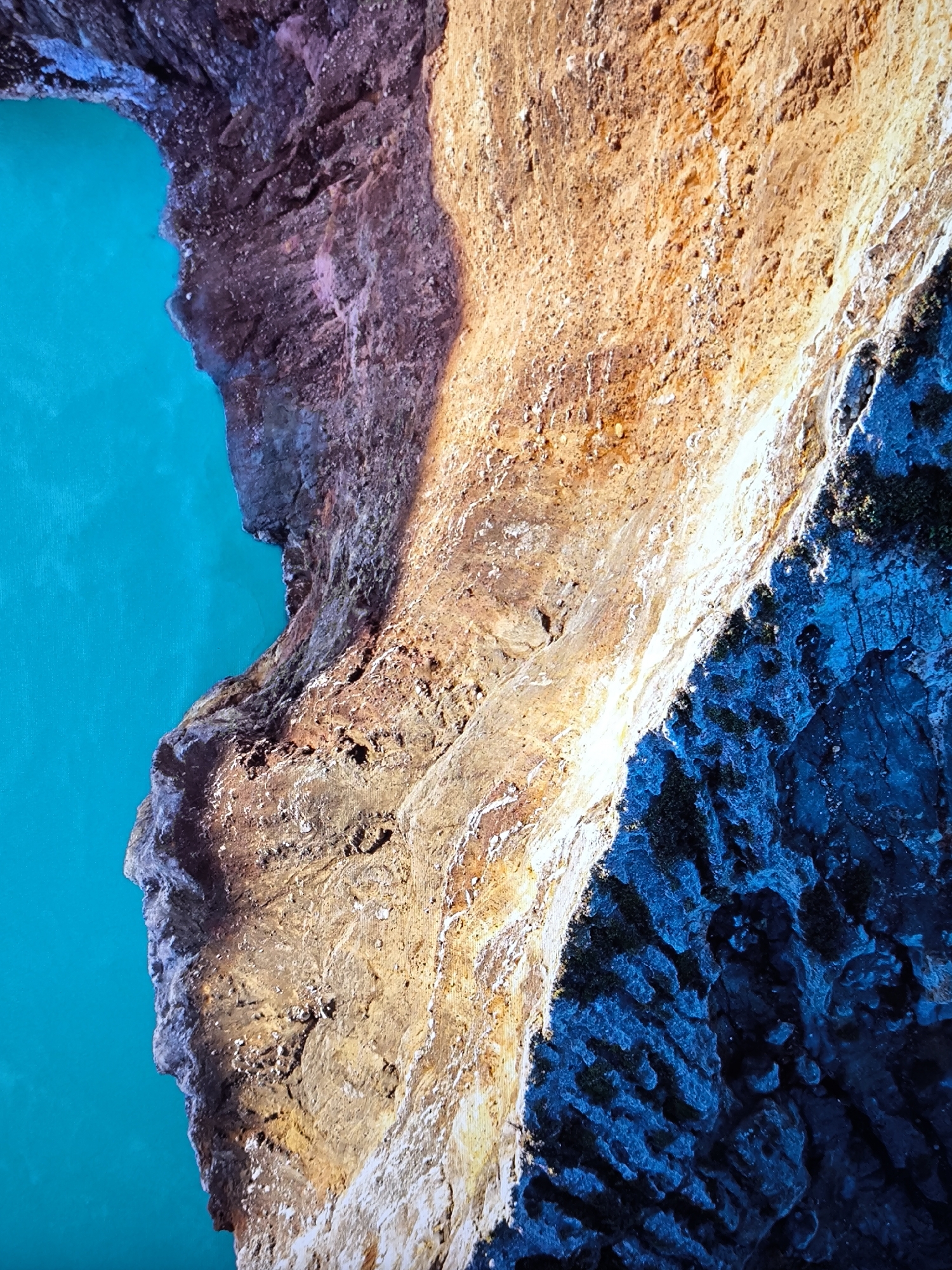 The image displays an aerial or high-angle perspective of a natural geological formation at the city of Unknown, Unknown. A vivid turquoise body of water occupies approximately the left third of the frame, bordered by an irregular, rugged cliff face that extends across the central and right portions. The upper-central section of the cliff is brightly illuminated, presenting warm tones of orange, reddish-brown, and tan, with highly textured rock formations and visible erosion patterns. A sharp diagonal shadow line traverses the cliff from top-right to bottom-center, separating the sunlit areas from the lower-right portion, which is cast in deep blue and dark gray shadows, revealing complex rock structures and sparse, dark vegetation. The interface between the turquoise water and the cliff is a distinct, meandering waterline. No human subjects, animals, or man-made objects are present in the scene.