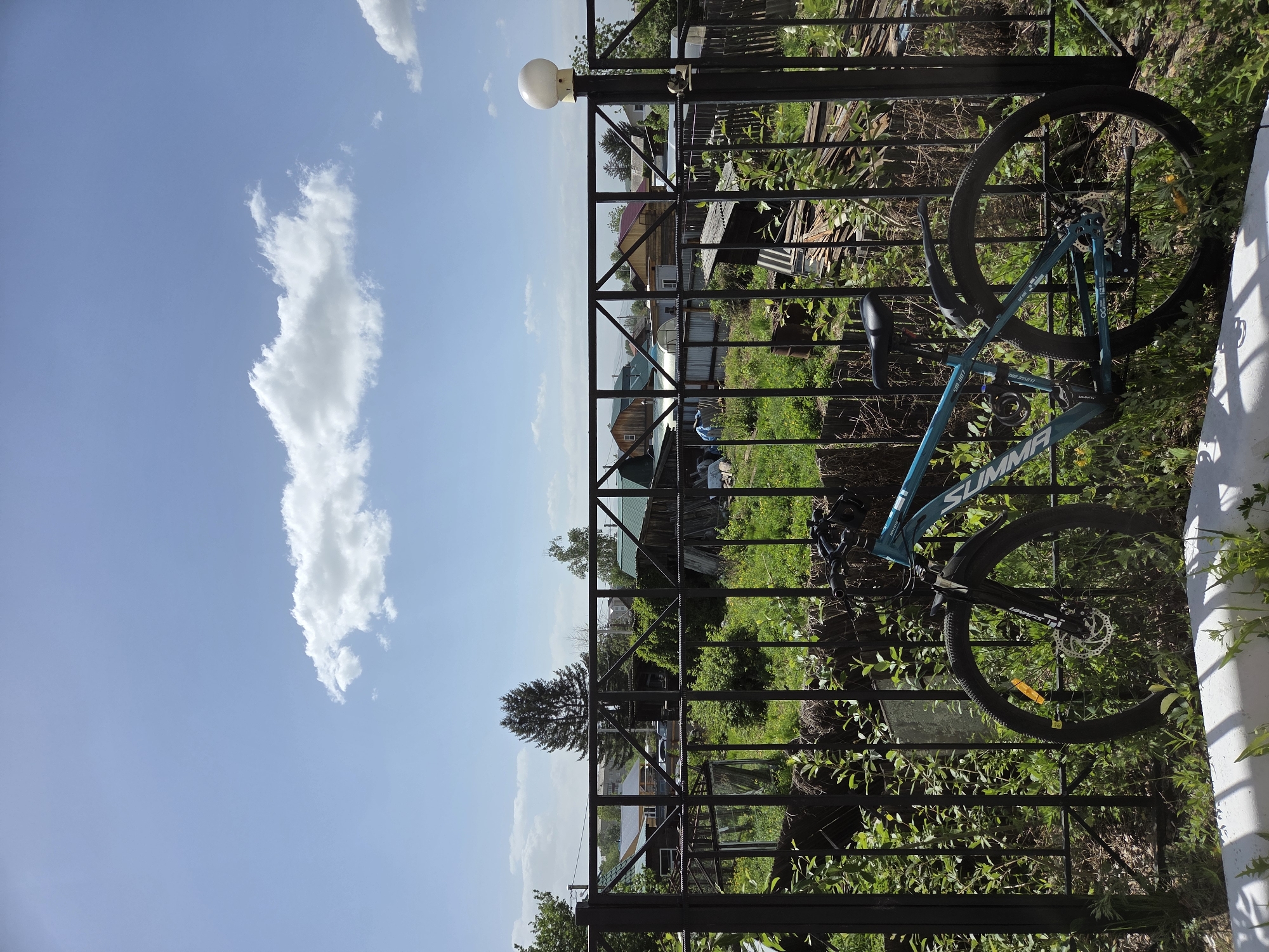 A teal bicycle rests against a black metal fence in an outdoor setting. Beyond the fence, there is lush green vegetation and several buildings, including houses with varying roof colors. The sky above is a clear blue with a prominent white cloud. The image appears to be taken from a slightly low angle, looking upwards towards the sky. The bike has its tires touching the ground, and the frame is leaning against the fence. The scene is well-lit with natural sunlight. Location details indicate this is Birobidzhan, Russia, though there are no specific details in the image to verify this.