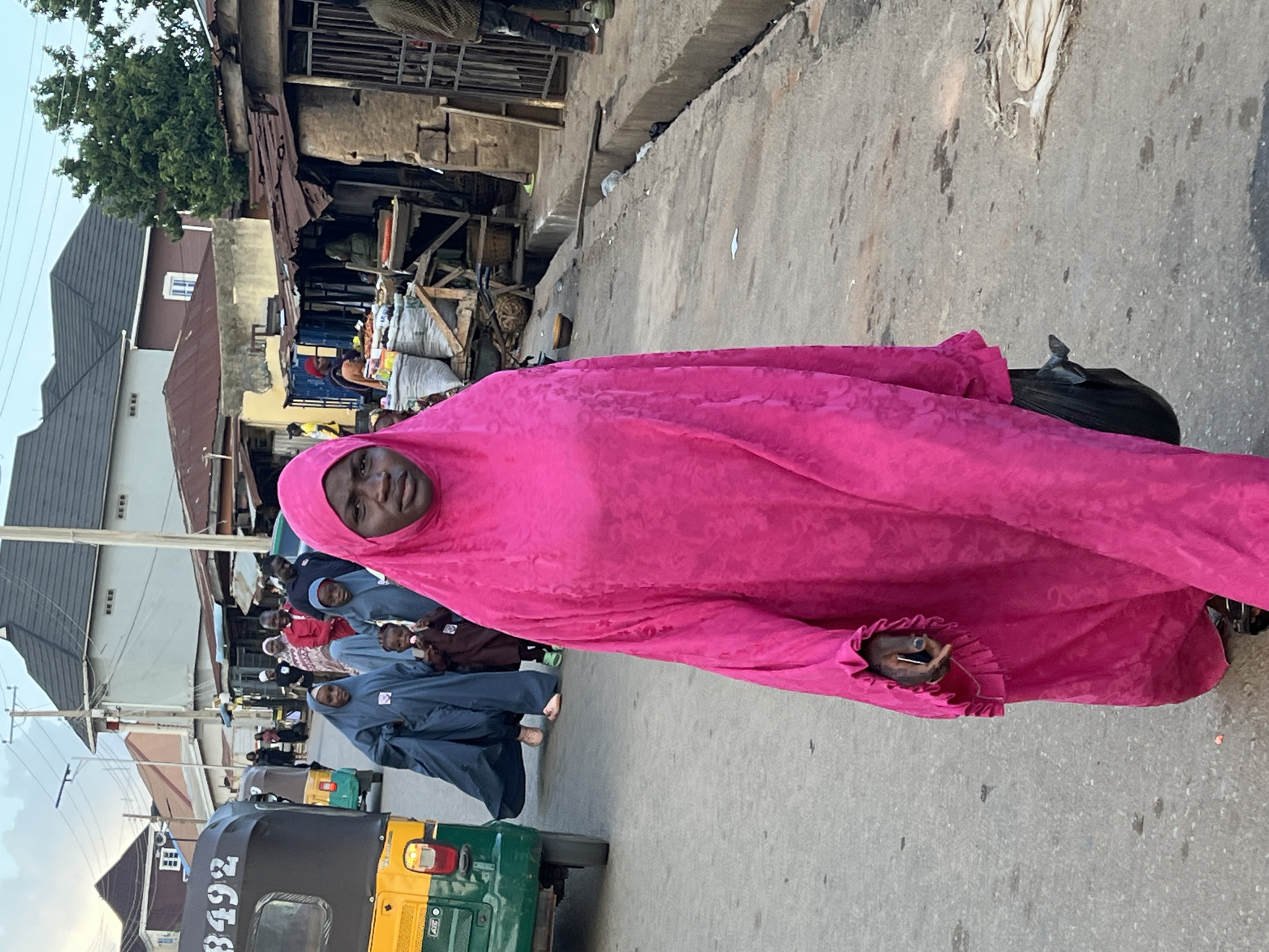 An outdoor street scene in Jos, Nigeria, captures a woman in a vibrant pink hijab walking towards the camera, holding a black plastic bag in her left hand. She is the primary subject in the foreground. Behind her, a group of people are visible, including several women and children in blue and brown hijabs. To the left, a yellow and green tricycle, a common mode of transport in Nigeria, is parked. The background depicts a street lined with buildings, some with corrugated iron roofs, and market stalls with goods displayed. The environment suggests a bustling urban setting.
