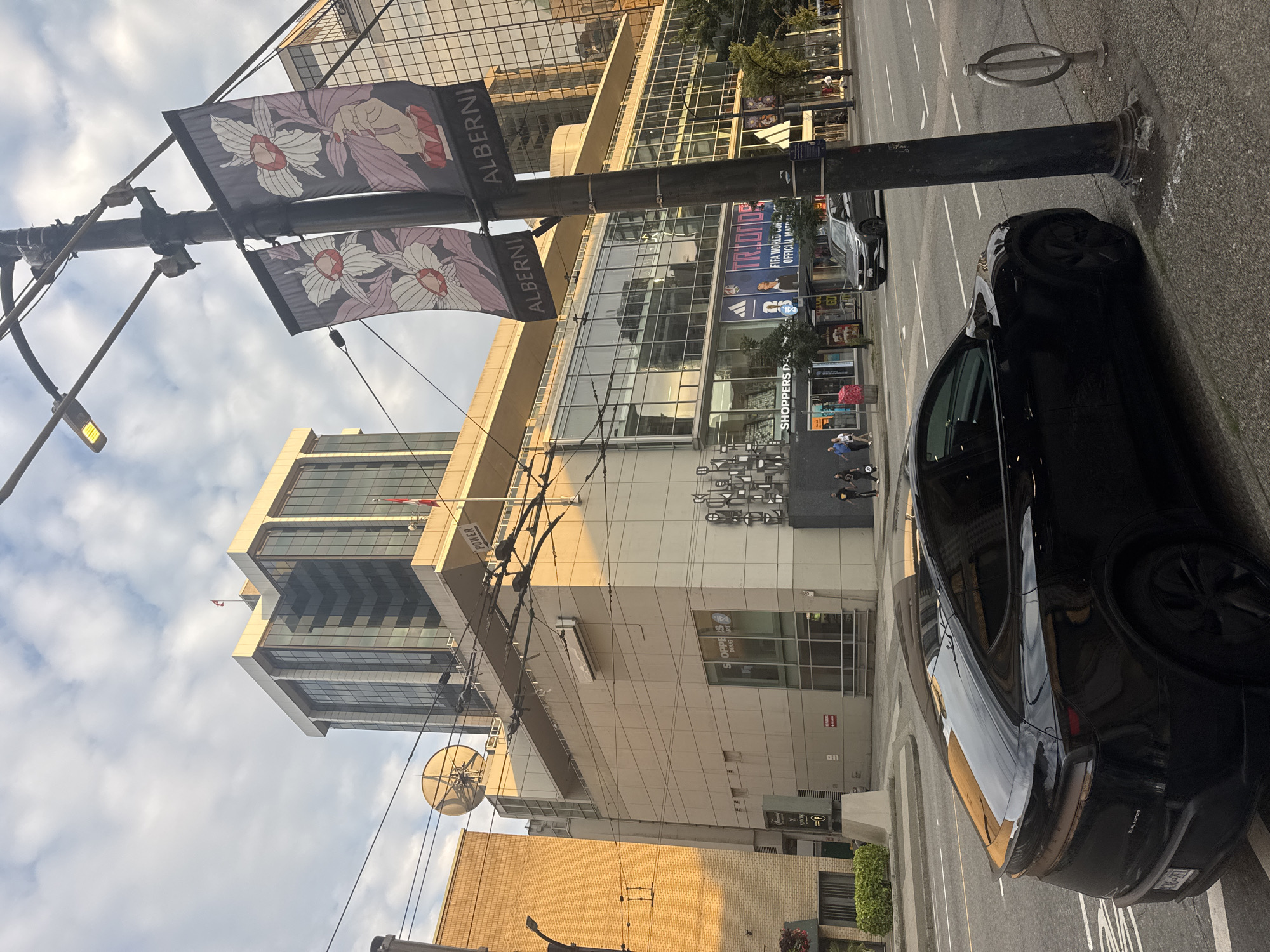 The image depicts a street scene in Vancouver, Canada, likely on Alberni Street, as indicated by banners on a lamppost. In the foreground, a black electric SUV is parked at the curb. The vehicle is facing right, with its rear visible. Across the street, a multi-story building houses various businesses, including "Shoppers Drug Mart" and "Trionfo". The architecture of the building features large glass windows reflecting the sky and surrounding buildings. Above the street level, there are overhead power lines and traffic signals. In the background, modern high-rise buildings are visible, some with glass facades. The sky is partly cloudy with sunlight illuminating parts of the scene, suggesting it is either morning or late afternoon. A few pedestrians are walking on the sidewalk.