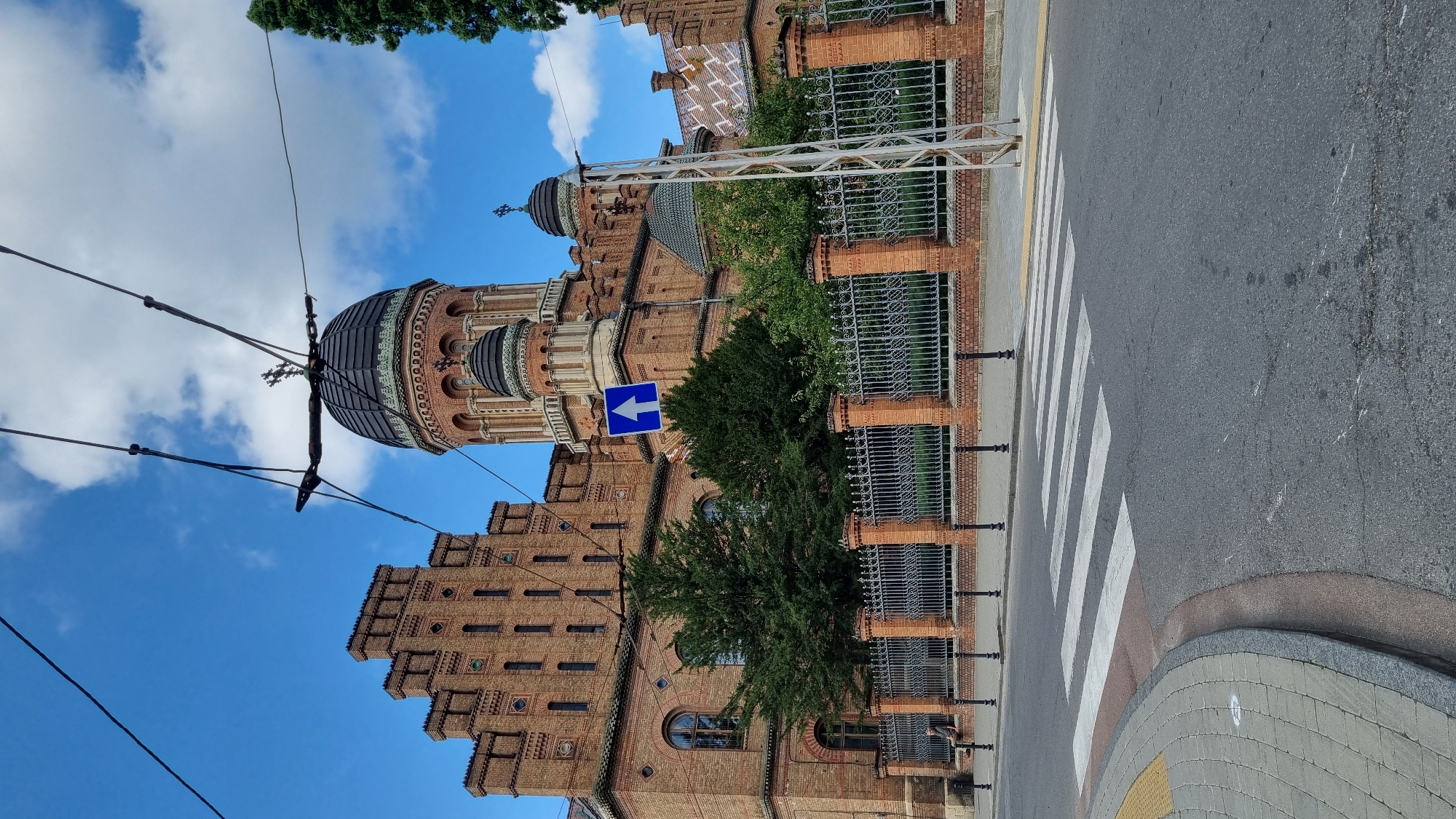 The image depicts the Chernivtsi National University named after Yuri Fedkovych in Chernivtsi, Ukraine. The grand architectural complex, constructed primarily of red brick, features intricate detailing and multiple domes. In the foreground, a pedestrian crossing with white stripes is visible on an asphalt road. A curved sidewalk with grey paving stones and yellow accents borders the crossing. A wrought-iron fence with brick pillars separates the university grounds from the street. Lush green trees are planted behind the fence. Overhead, power lines and a blue traffic sign with a white arrow pointing upwards are present. The sky is blue with scattered white clouds. A single person is seated on a low wall to the left of the image, outside the fence. The overall scene is an exterior view of the university's iconic buildings and surrounding urban environment under daylight.