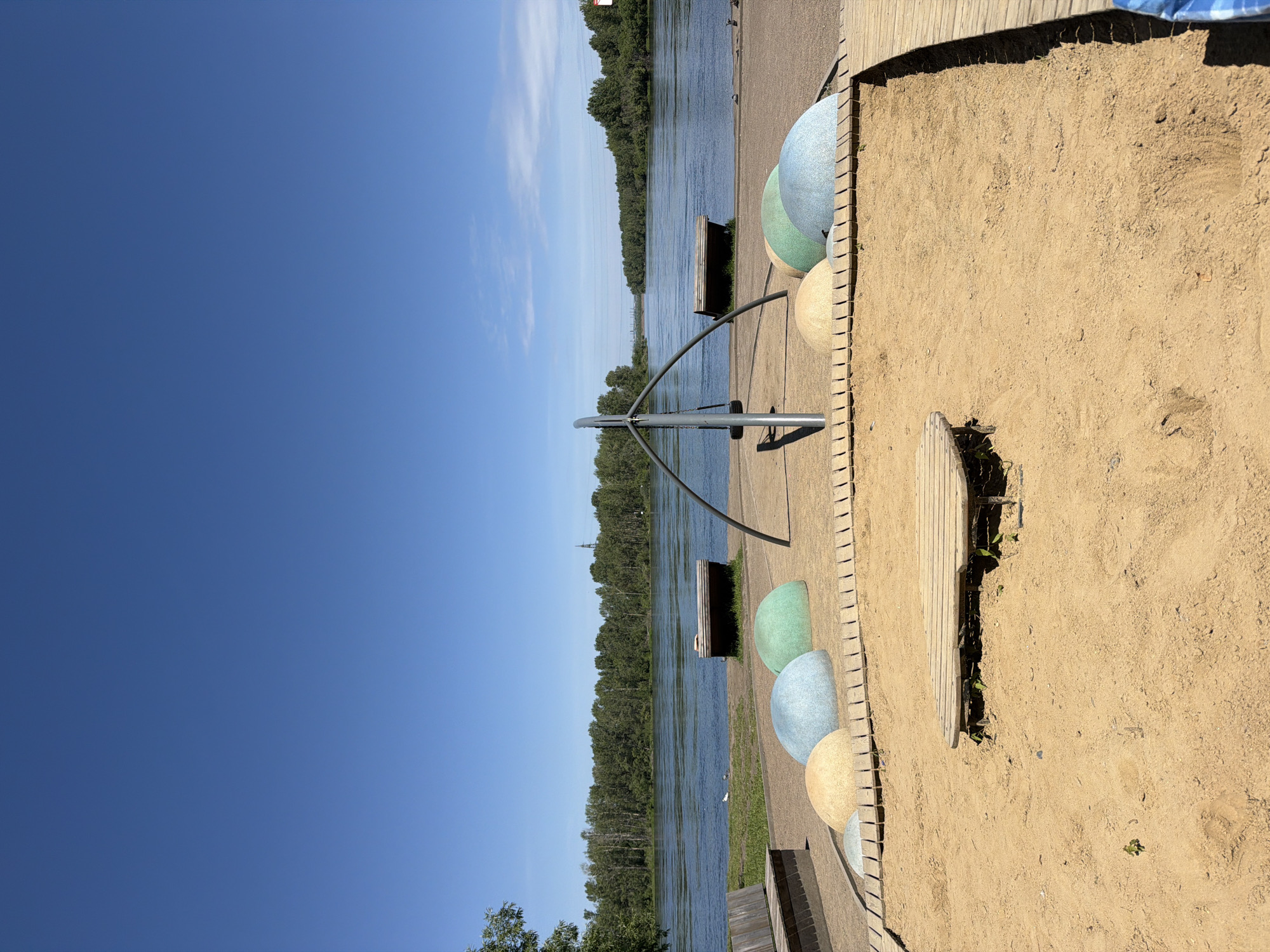 This is a vertical, eye-level shot of a beach area next to a wide, calm river under a clear blue sky with a few wispy clouds. The sun is bright, suggesting it is daytime, likely late morning or afternoon. The beach is composed of light brown sand with several large, rounded, colorful decorative objects scattered on it, in shades of light blue, green, and yellow. Some of these objects are partially buried in the sand. A wooden boardwalk runs along the edge of the sand.
In the background, the river stretches into the distance, lined by a dense forest of green trees. The water reflects the sky and trees. A wooden bench is visible near the water's edge. A modern, minimalist metal sculpture with curved supports is also present on the sand, near the river. The overall scene appears to be a recreational area, possibly a park or a waterfront attraction, designed for relaxation and enjoyment in a natural setting. There are no people visible in the image, and no text is present. The mood is serene and peaceful. The location context is Krasnoyarsk, Russia.