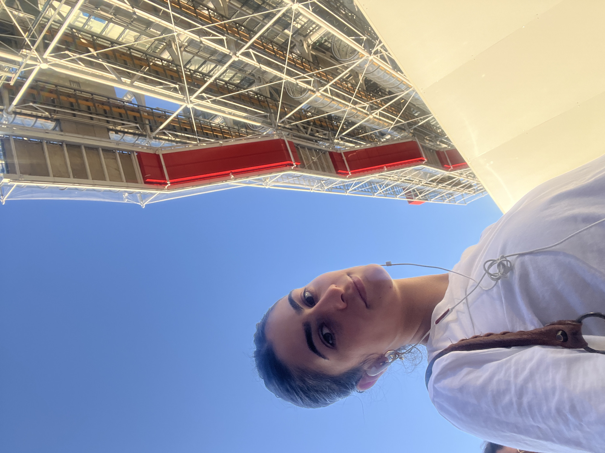 A low-angle shot captures a woman's face against a clear blue sky, with a building structure in the background. The woman has dark hair, fair skin, and is wearing a white shirt with earphones. The building features a complex network of metal beams and what appears to be ductwork. Red elements are visible on the building's exterior. The location is Paris, France. The building is the Centre Pompidou.