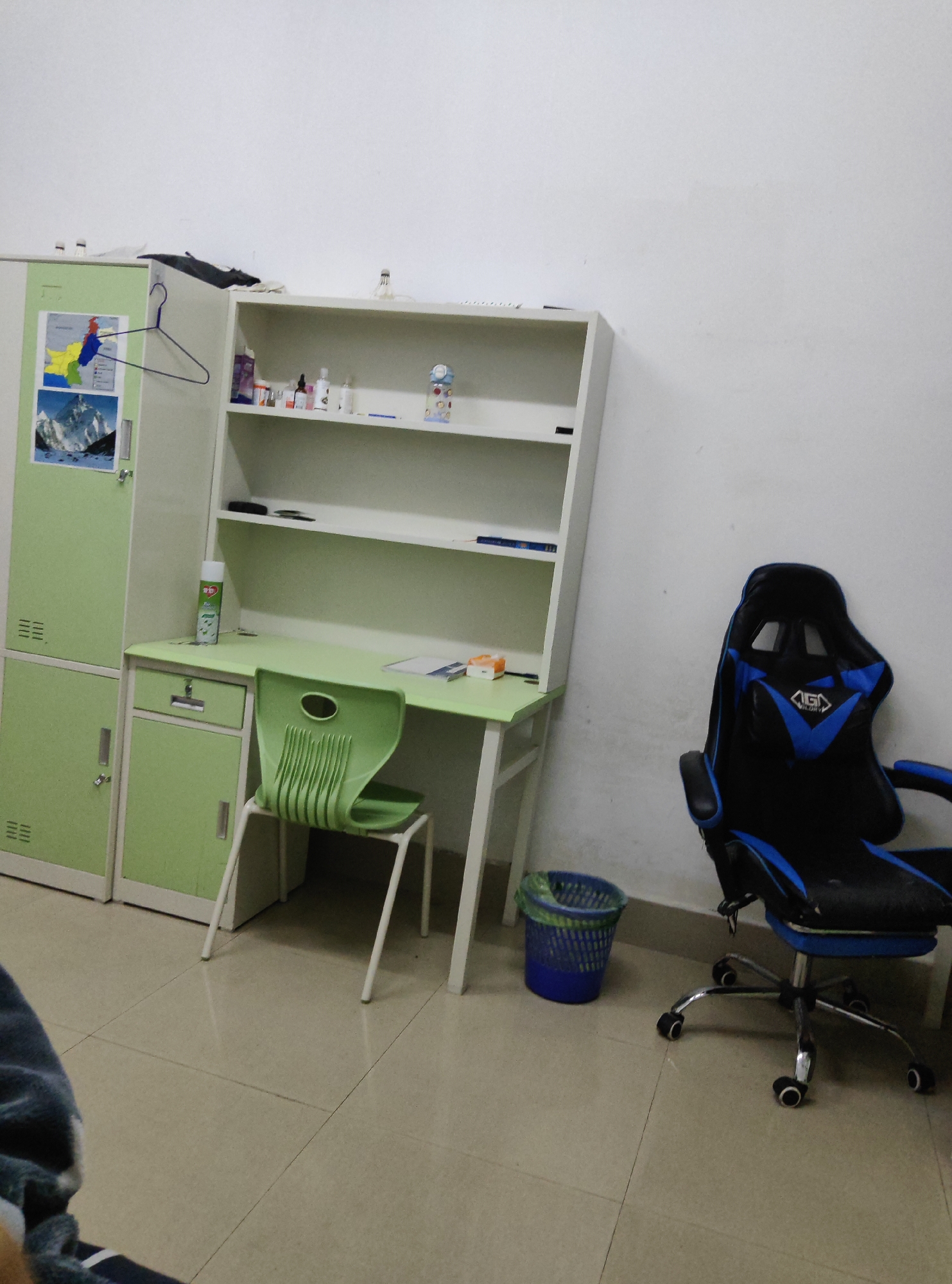 An indoor scene depicts a room that appears to be a student's dorm or a small office. The room is furnished with a light green metal locker unit on the left, a matching desk with a light green surface and drawers, and a white shelf unit above the desk. A green plastic chair is positioned at the desk. A blue and black gaming chair with wheels is situated to the right of the desk, facing away from it. A blue mesh trash can with a green liner is on the floor near the desk. The walls are painted white, and the floor is tiled with beige tiles. The scene is likely located in the city of Chongqing High-tech Zone, China, as indicated by the presence of these common types of furnishings in student dormitories or modern office spaces within such areas. No people are visible in the image, and no specific actions or interactions are taking place.