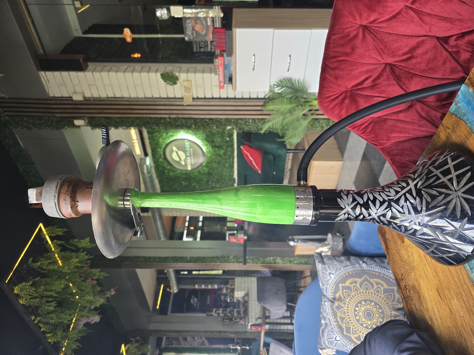 The image displays a close-up view of a modern hookah (shisha) situated on a wooden table with a blue resin inlay, likely in a lounge or cafe in Sofia, Bulgaria. The hookah itself features a vibrant light green glass stem, a dark base intricately patterned with silver leaf or star designs, a metal tray, and a brown ceramic bowl on top. A black hose extends from the side of the hookah.
Surrounding the hookah are various elements indicating a comfortable and stylish setting. To the right, a plush red velvet tufted chair or cushion provides seating. On the left, a blue cushion adorned with a golden and grey mandala pattern is visible, suggesting additional seating.
The background features a prominent green artificial hedge wall, against which a circular illuminated sign is mounted. This sign clearly reads "HOOKA PLANET" with a stylized planet graphic, and "SHISHA CLUB" underneath, identifying the establishment. Further into the background, a green velvet chair with a distinctive red lip-shaped cushion is visible. Artificial plants, including ferns and other greenery, are placed around the space, enhancing the lounge's decor. Reflections on glass surfaces in the background show partial text like "brunch" and "menu 9:00", as well as a "WI-FI" sign. Yellow string lights draped over plants in the distance suggest an evening or nighttime setting, creating a cozy and inviting ambiance.