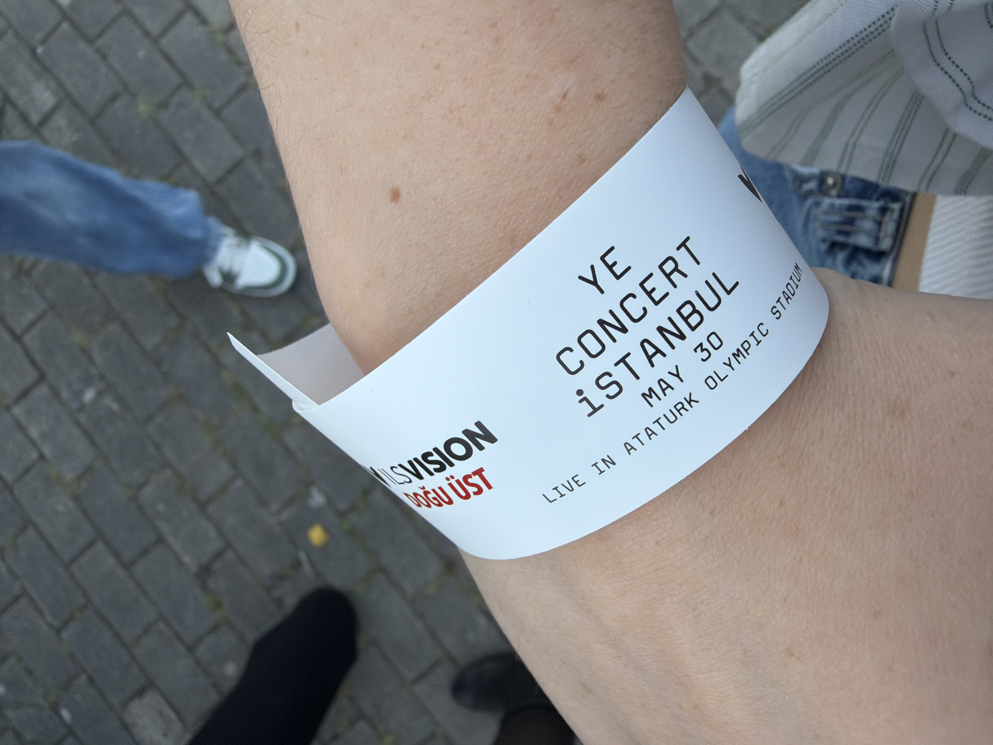A person is wearing a white wristband on their right wrist. The wristband reads "YE CONCERT İSTANBUL MAY 30 LIVE IN ATATURK OLYMPIC STADIUM". The wristband also has "MİLSVİSİON DOĞU ÜST" printed on it. The person is standing outside on a brick surface, with other people's legs and shoes visible in the background. The location is the Atatürk Olympic Stadium in Başakşehir, Turkey.