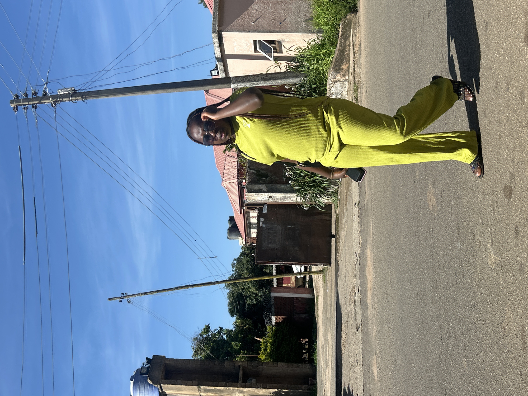 A young Black woman with braided hair and sunglasses walks on a paved road under a clear blue sky in Jos, Nigeria. She is wearing a lime green, ribbed two-piece set, consisting of a short-sleeved top and wide-leg pants. She holds a dark-colored mobile phone in her left hand and adjusts her sunglasses with her right hand. She is wearing dark sandals. In the background, there are buildings with red roofs, a tall utility pole with many wires, and lush green trees. The road appears to be a residential area.
