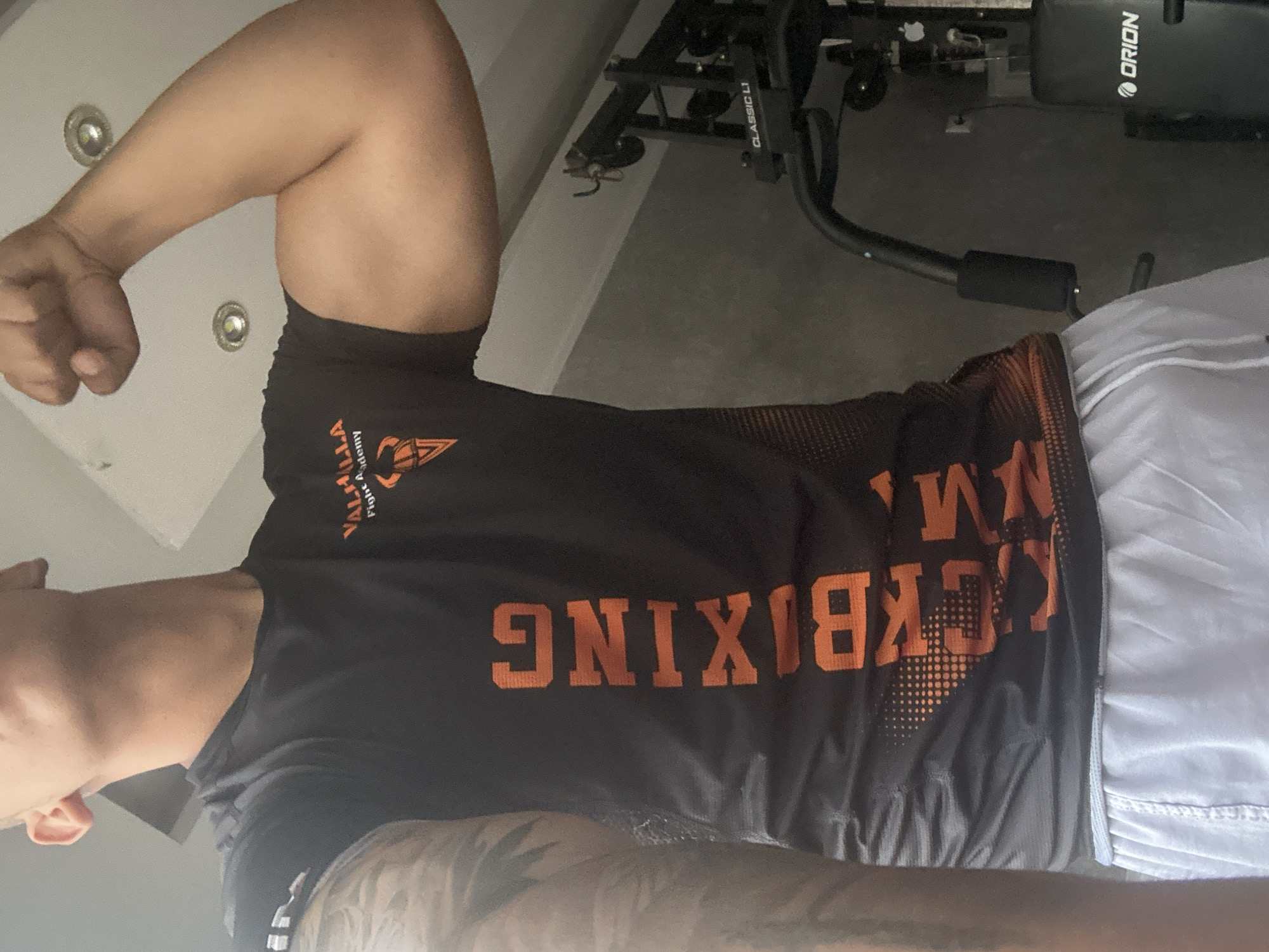 A man is flexing his arm while wearing a black t-shirt with orange lettering that reads "Kickboxing." The t-shirt also displays a logo, partially obscured by the arm. The man is wearing grey shorts. In the background, a piece of exercise equipment labeled "Orion" and "Classic L1" is visible. The location appears to be indoors, possibly a home gym.