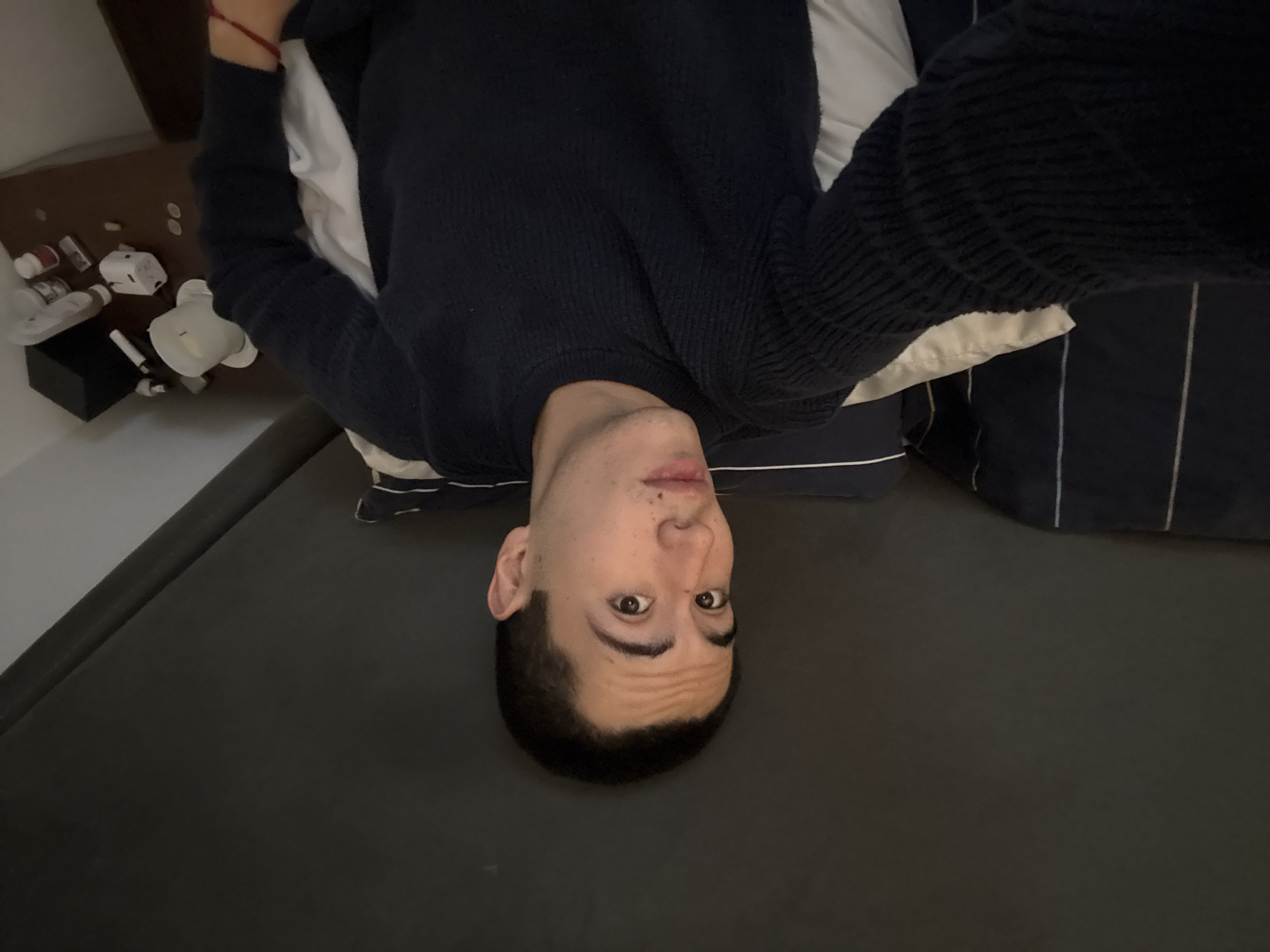 A selfie of a person with fair skin, dark hair, and brown eyes is taken from an upside-down perspective. The person is wearing a dark blue sweater and lying on a dark grey or black surface, likely a bed. White pillows are partially visible behind their head. In the upper left corner of the frame, there is a bedside table with various objects, including white bottles, a small black box, and a white figurine. The photo does not contain enough information to determine if it was taken in San Bernardino Tlaxcalancingo, Mexico.