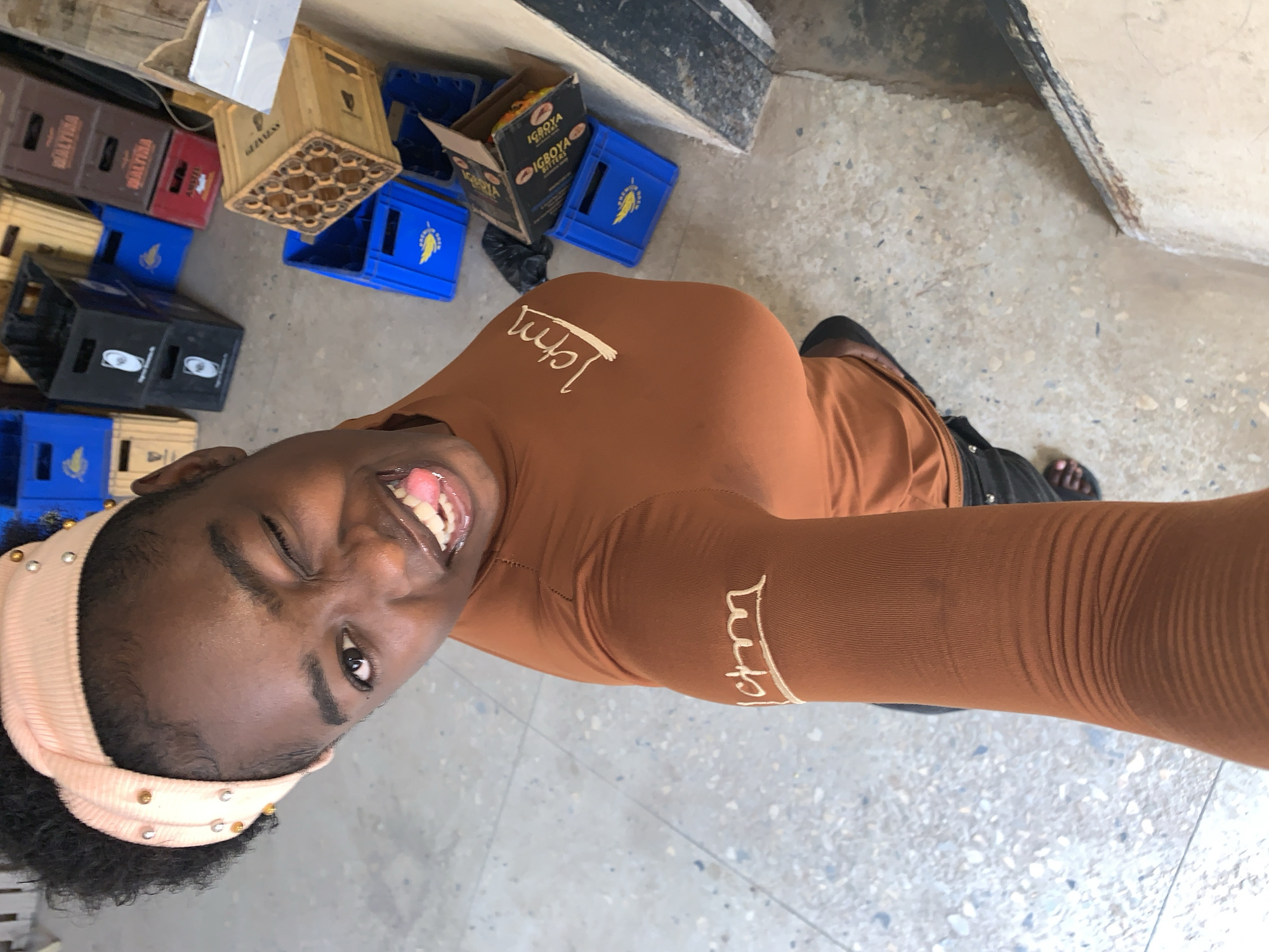 A young Black woman with short dark hair and a peach-colored headband with small metallic studs is taking a selfie in what appears to be a warehouse-type setting. She is winking with her right eye and sticking her tongue out, smiling widely. She is wearing a long-sleeved brown top with the word "Jam" embroidered on the chest and arm in a lighter brown or beige thread.
Behind her and to the left, there are stacks of various colored plastic crates, including brown, red, yellow, black, and blue. Some wooden crates are also visible. A cardboard box with the words "IGBOYA Bitters" is visible amongst the crates.
The floor is a light grey with speckled texture. The background includes parts of a light-colored wall and a darker grey or black structure near the top of the frame. The location is Kabong, Nigeria.