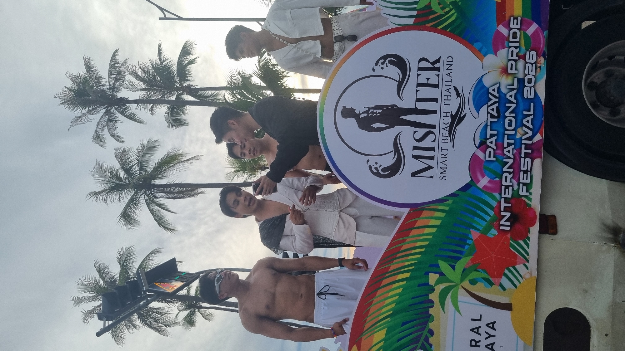 The image shows a group of young men participating in a pride festival parade. The setting appears to be outdoors, possibly on a street lined with palm trees, and the sky is overcast, suggesting it might be late afternoon or early morning. The participants are diverse in their attire, with some wearing white outfits that appear to be part of a costume, while others are shirtless or in casual wear. One individual in the foreground is shirtless, wearing white shorts and sunglasses. Another man, partially visible behind him, is also shirtless. Further back, a group of men are gathered around a large circular banner.
The banner is a prominent object in the image, featuring vibrant rainbow colors and various graphics. Text on the banner reads "MISTER SMART BEACH THAILAND" within a circular logo that depicts a silhouette of a muscular man. Below this, more text states "PATTAYA INTERNATIONAL PRIDE FESTIVAL 2025". The overall mood seems festive and celebratory, with the participants appearing engaged and in good spirits. The presence of palm trees and the text "Pattaya" strongly indicate the location as Pattaya City, Thailand. The event itself is clearly a pride festival, likely a parade or street celebration, given the presence of the banner and the gathering of people.