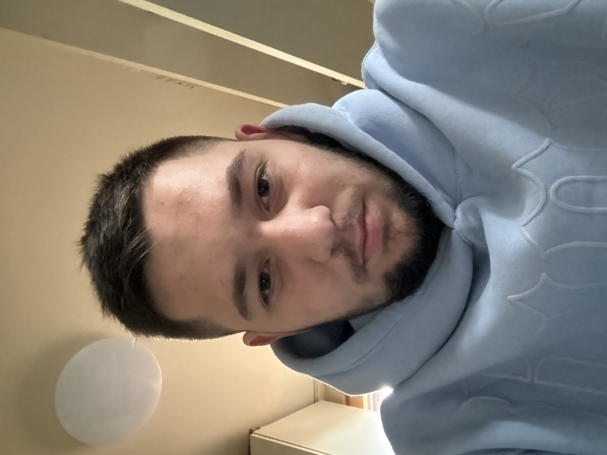 A portrait of a man with short dark hair, brown eyes, and a dark beard is the primary subject. He is wearing a light blue hoodie with a raised pattern on the fabric. The background includes a light-colored wall and ceiling. There is a white round object affixed to the wall, possibly a light fixture. A white cabinet or piece of furniture is visible. There is no indication of the location of Sofia, Bulgaria in the image.