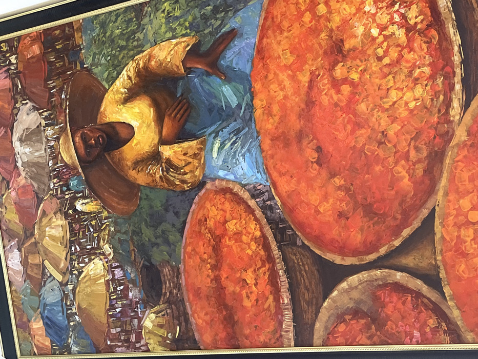 This oil painting depicts a vibrant outdoor market scene located in the city of Alaguntan, Nigeria. In the foreground, a dark-skinned individual, wearing a broad-brimmed straw hat, a bright yellow long-sleeved tunic, and a blue lower garment, is seated with a direct gaze forward. Their right hand rests on their lap, while the left hand is extended to the side. Positioned prominently around this figure are several large, round, woven baskets filled with a copious amount of vivid orange-red granular material, consistent with a foodstuff such as dried peppers or spices. The mid-ground and background reveal a bustling market ambiance characterized by numerous colorful umbrellas and canopies in shades of red, orange, brown, blue, and white, providing shade over indistinct market stalls and numerous background figures. The ground beneath the market appears to be a mix of earthy tones and green elements, indicative of an open-air setting. The entire composition is rendered with thick, visible impasto brushstrokes.