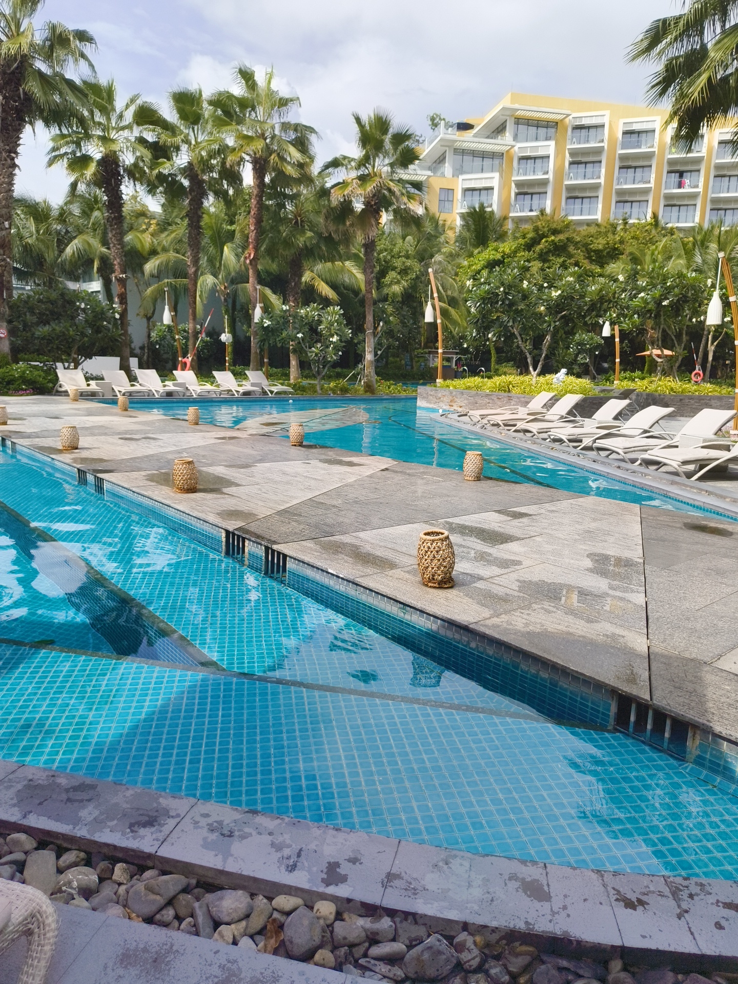 The image captures a resort swimming pool area on a cloudy day in Phú Quốc, Vietnam. The foreground features a turquoise tiled swimming pool with a complex geometric design on the tiled floor. Adjacent to the pool is a paved area with large, grey stone tiles, some of which appear wet. Scattered across this paved area are several woven basket-like lanterns.
Beyond the paved area, a row of light-colored lounge chairs are neatly arranged by the pool's edge. Behind the lounge chairs, a dense line of palm trees and other lush green foliage separates the pool area from a tall, modern hotel building. The hotel is painted a bright yellow and features multiple stories with balconies and large windows. The sky above is overcast, with patches of white clouds visible. The overall impression is one of a well-maintained resort environment designed for relaxation and leisure. No people are visible in the frame.
