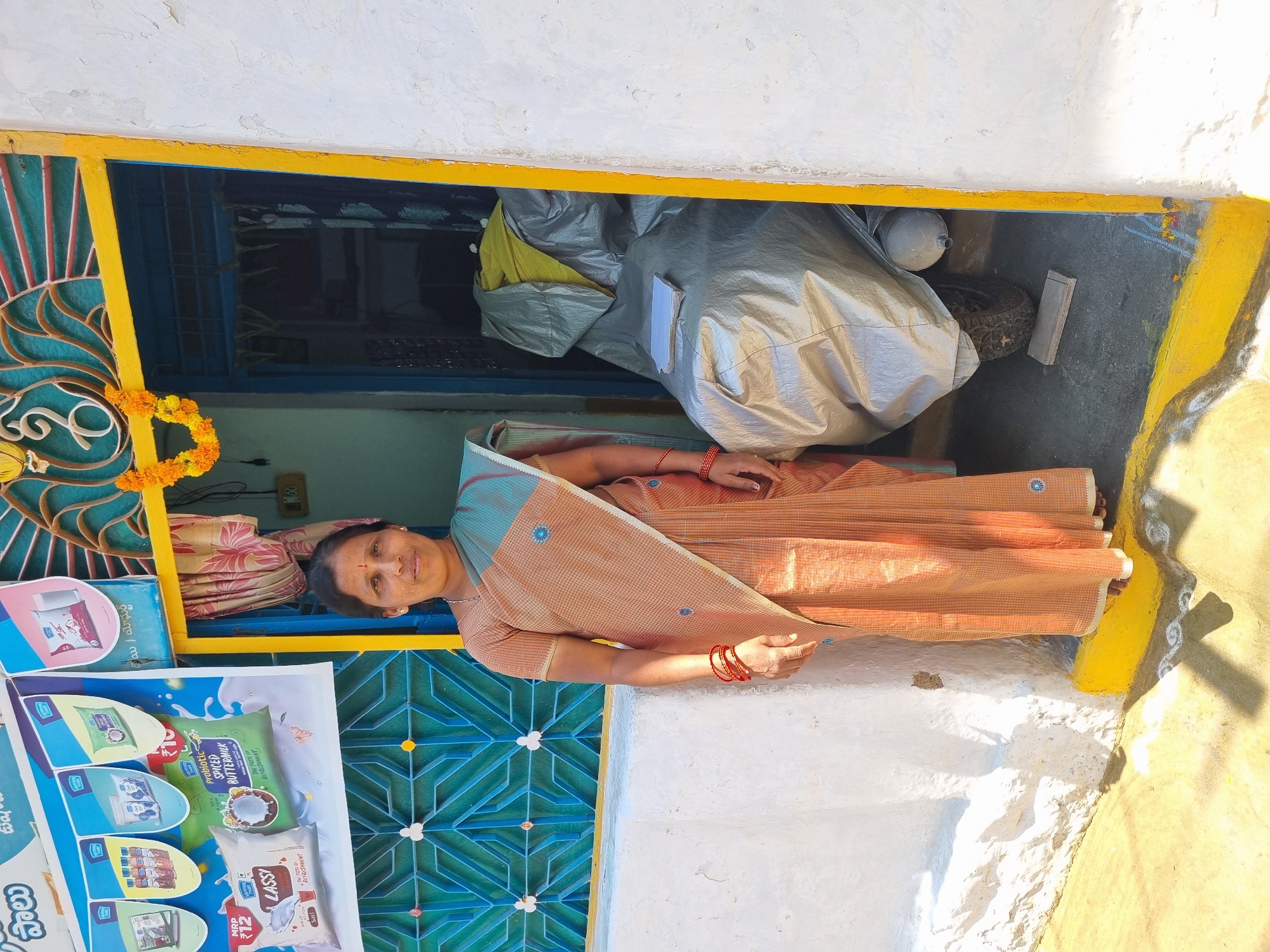 A full-length, front-facing image shows an adult Indian woman standing in a sunlit doorway in Pedda Kammavaripalli, India. The woman, appearing to be in her mid-forties, wears a light peach-colored striped saree with blue accents and small blue circular motifs, draped over a matching short-sleeved blouse. She has red bangles on both wrists, a small red bindi on her forehead, and a subtle smile. Her dark hair is pulled back.
To her left, a white textured wall features a light blue window covered by a geometric pattern grill. Above and partially obscuring the window, a multi-colored banner advertises "Hatsun Daily" dairy products, including Lassi and Buttermilk, with product images, Telugu text, and "MRP ₹12" visible.
The doorway is framed by a bright yellow outer border and a blue inner frame. Above the blue frame, an ornate teal and red metal grill is visible, beneath which a marigold garland hangs. Inside the doorway, a motorcycle or scooter is mostly covered by a silver tarpaulin, with a stack of white papers resting on top. A yellow fabric is visible near the top of the covered vehicle. A black tire and an exhaust pipe are partially visible beneath the cover, with a small wooden block on the dark concrete floor.
To the woman's right is a continuation of the white textured wall. The ground directly outside the doorway is light-colored concrete or packed earth, bordered by a yellow stripe at the base of the wall, with a faint white rangoli pattern visible. The scene is brightly lit by direct sunlight, casting distinct shadows.