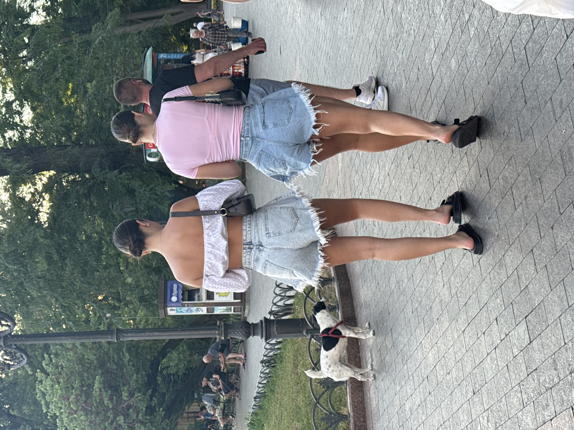 Two young women wearing denim shorts and crop tops walk away from the camera on a paved walkway in an urban park in Odesa, Ukraine. The woman on the left wears a white off-the-shoulder top, and the woman on the right wears a pink t-shirt. Both women have their hair pulled back in buns. A white and black dog with a red harness stands to the left of the women, facing them. In the background, several people sit on park benches, and trees line the park. A lamppost is visible to the left of the women. The overall atmosphere is casual and suggests a warm day in the city.