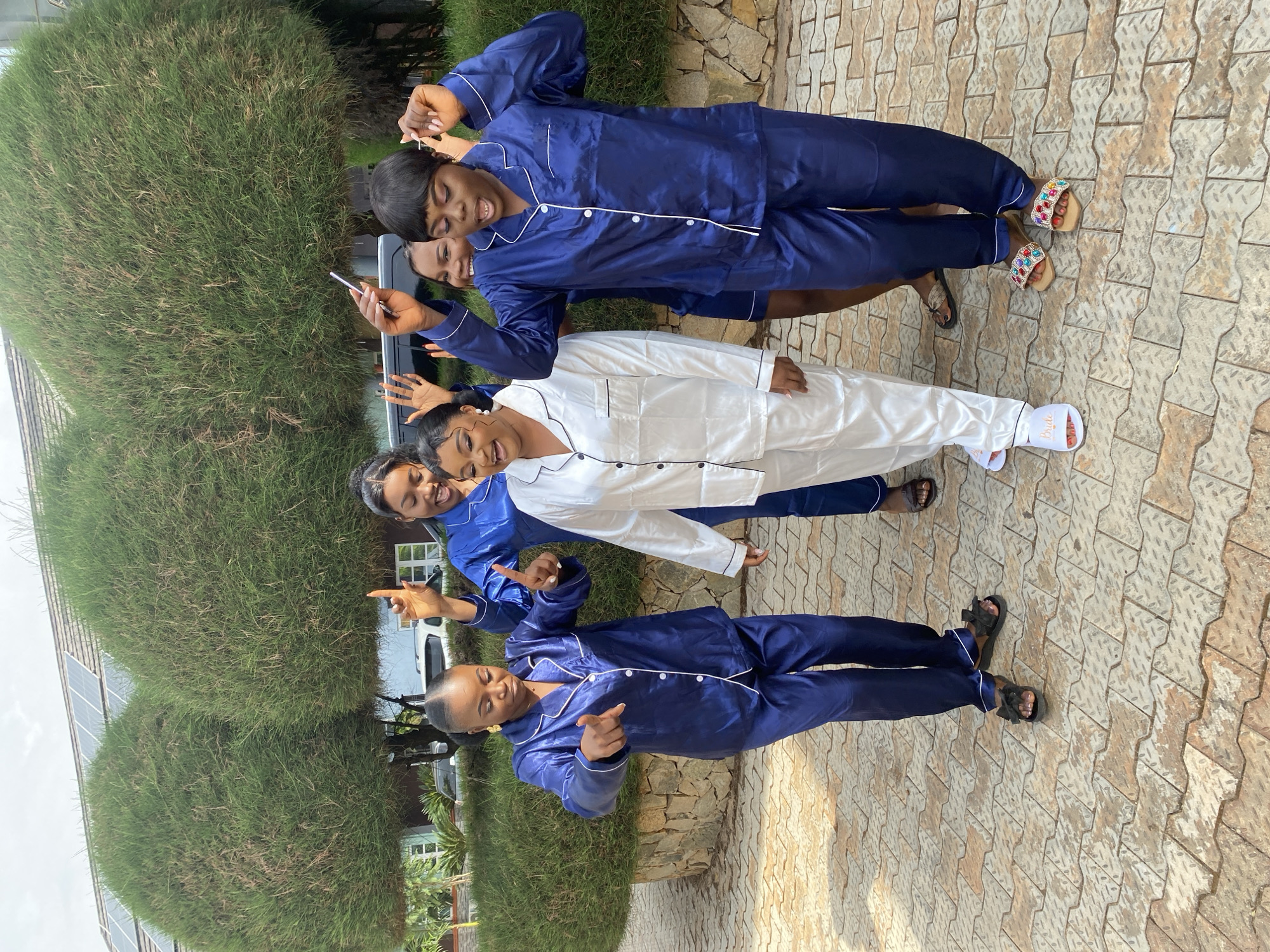 Four women are posing in a line outside, wearing pajamas. Three women are wearing navy blue pajamas with white piping, and one woman is wearing white pajamas with black piping. One woman in blue is holding a smartphone. They are standing on a paved area that has a brown diamond pattern. Behind them are green bushes. The location appears to be Bar Jirgi Summa, Nigeria.