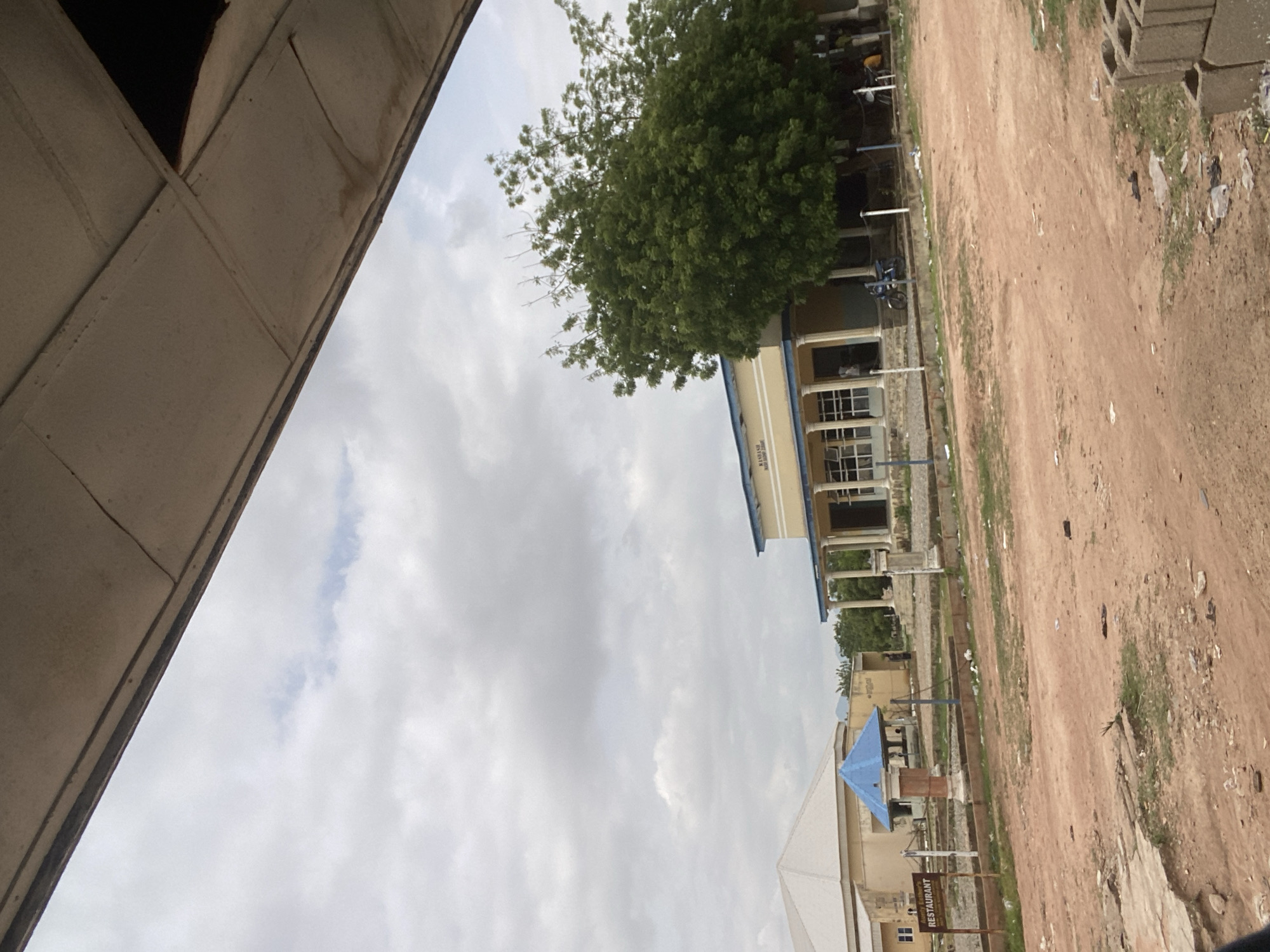 The image depicts a building with a light yellow facade and a light blue trim, featuring a covered walkway supported by columns. A large, dense green tree stands to the right of the building, partially obscuring it. In the foreground, there is a dirt road with scattered debris. To the left of the main building, another structure with a light-colored, peaked roof is visible, along with a small, blue-canopied gazebo-like structure. A sign to the far left of the image reads "Aunty Esther's RESTAURANT". The sky is overcast with scattered clouds. The scene appears to be in Jalingo, Nigeria.