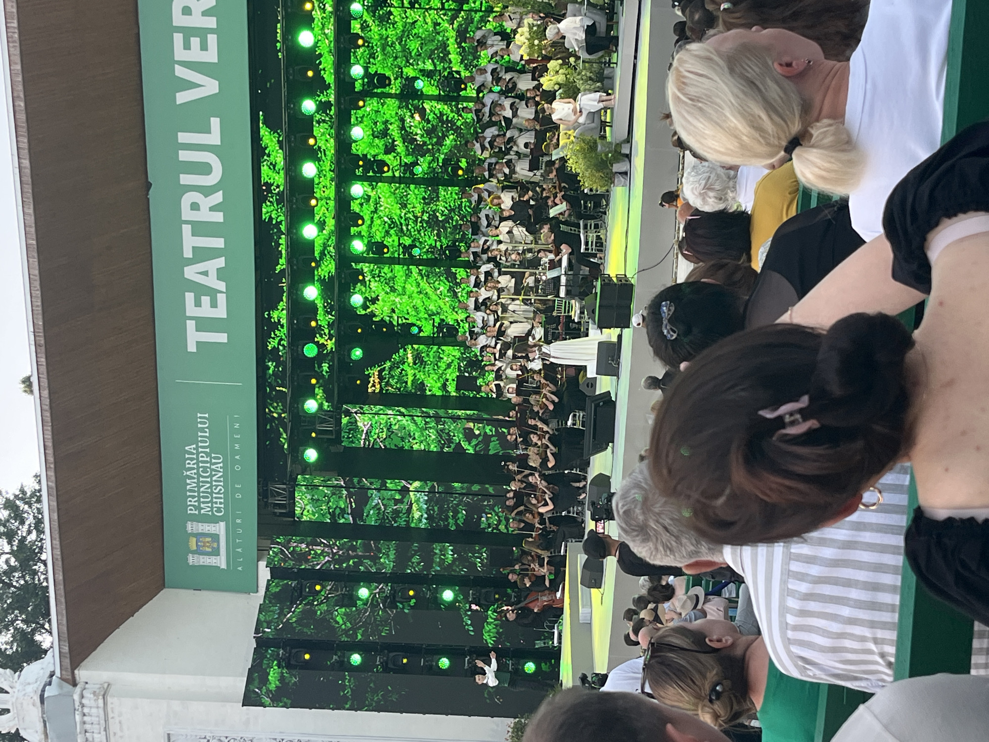The image depicts an outdoor concert at the "Teatrul Verde" (Green Theatre) in Chișinău, Moldova. A large orchestra and choir are performing on a brightly lit stage, which is decorated with a backdrop of lush green foliage. In the foreground, an audience is seated on green benches, facing the stage. Several members of the audience are visible, including a woman with blonde hair tied in a ponytail, a person with gray hair and a striped shirt, and a person with dark hair and a black top. The stage banner above the performance displays the text "PRIMĂRIA MUNICIPIULUI CHIȘINĂU" (Chisinau Municipality Mayor's Office) and "TEATRUL VER" (Green Theatre). The overall atmosphere appears to be one of a cultural event or festival.