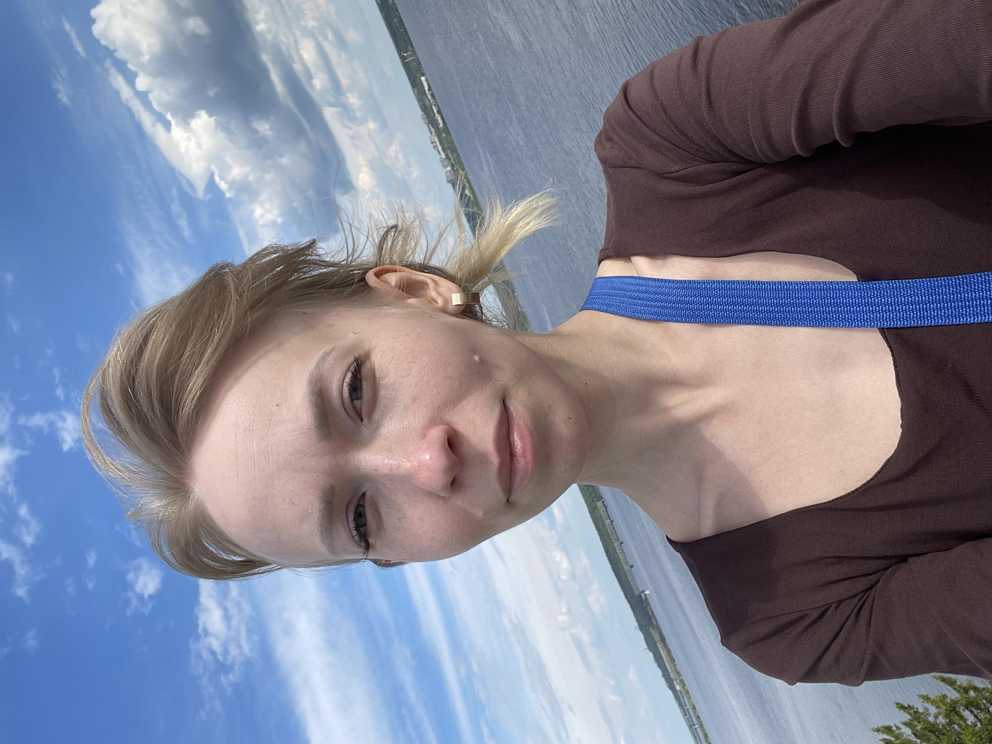 A selfie-style portrait captures a young adult female outdoors at what appears to be Пятый километр, Russia. The image is rotated 90 degrees clockwise, orienting the sky to the left and water to the right. The woman, with light skin and blonde hair tied back with some loose strands, looks directly at the camera with a neutral to slight smile. She has light-colored eyes and a few freckles or small marks visible on her face. She wears a dark brown long-sleeved top, and a prominent bright blue strap, possibly from a bag, crosses her chest. A gold-toned ear cuff is visible on her right ear.
The background is split between a clear blue sky with scattered white cumulus clouds on the left and a wide expanse of calm, dark blue water on the right. Along the distant horizon line across the water, faint traces of land with some structures or vegetation can be seen. A small portion of green foliage, likely a bush or tree, is visible in the bottom right corner of the frame. The bright lighting suggests it is daytime with sunny weather. There is no visible text in the image.