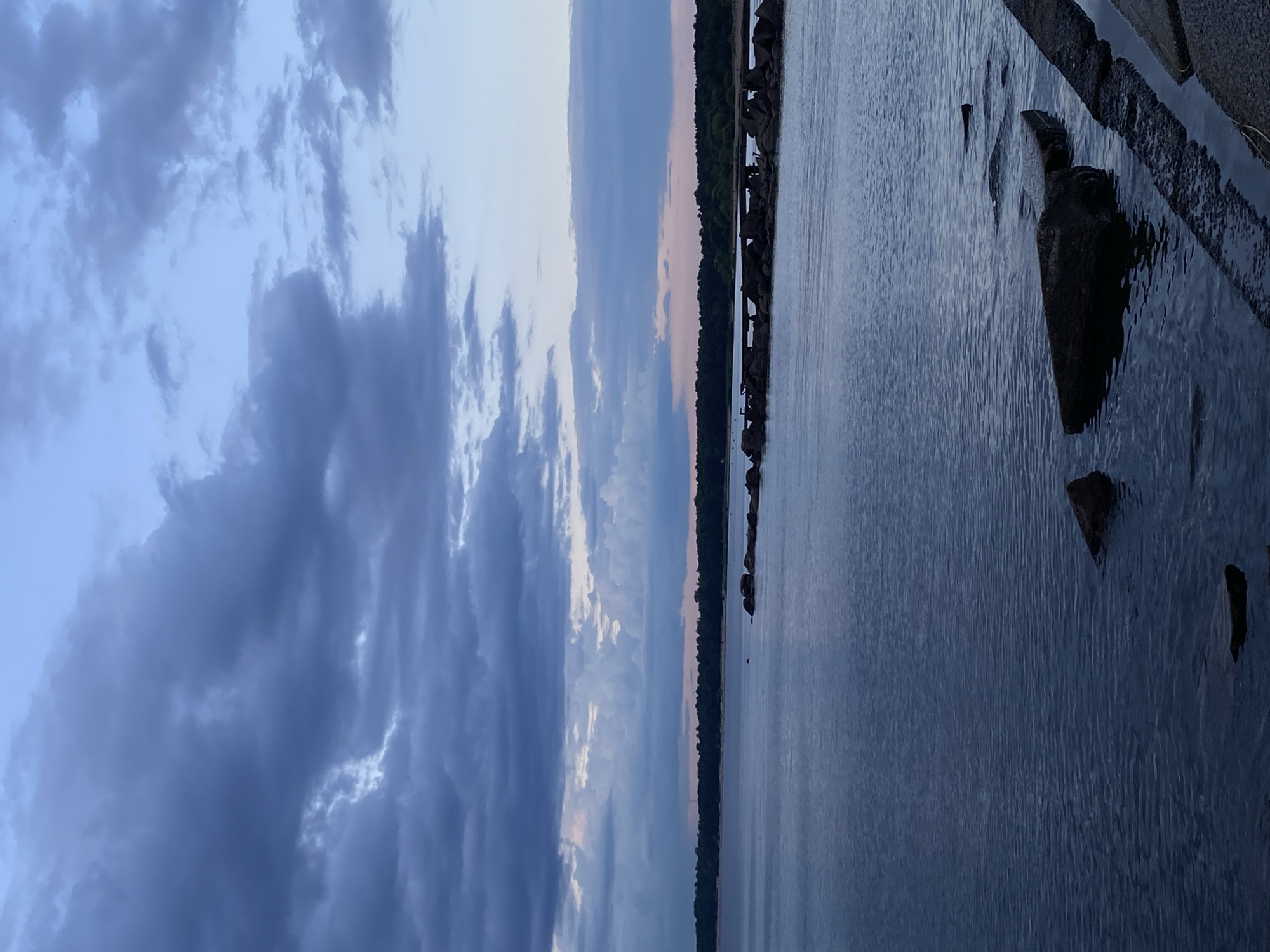 The image shows a calm body of water, likely the Gulf of Finland, near Sestroretsk, Russia. The water is dark blue and slightly rippled, reflecting the overcast sky. In the foreground, large rocks are visible partially submerged in the water and along the edge of a concrete embankment.
A line of larger rocks forms a breakwater or natural barrier in the middle ground, stretching horizontally across the image. Beyond the breakwater, a dense line of trees marks the distant shoreline. The sky above is a gradient of blues and grays, with heavy, dark clouds dominating the upper portion, transitioning to lighter, softer clouds closer to the horizon, tinged with pinkish hues. The overall mood is serene and tranquil, characteristic of twilight or an overcast day. There are no people or specific man-made structures clearly visible, besides the embankment in the foreground and the rocks in the water.