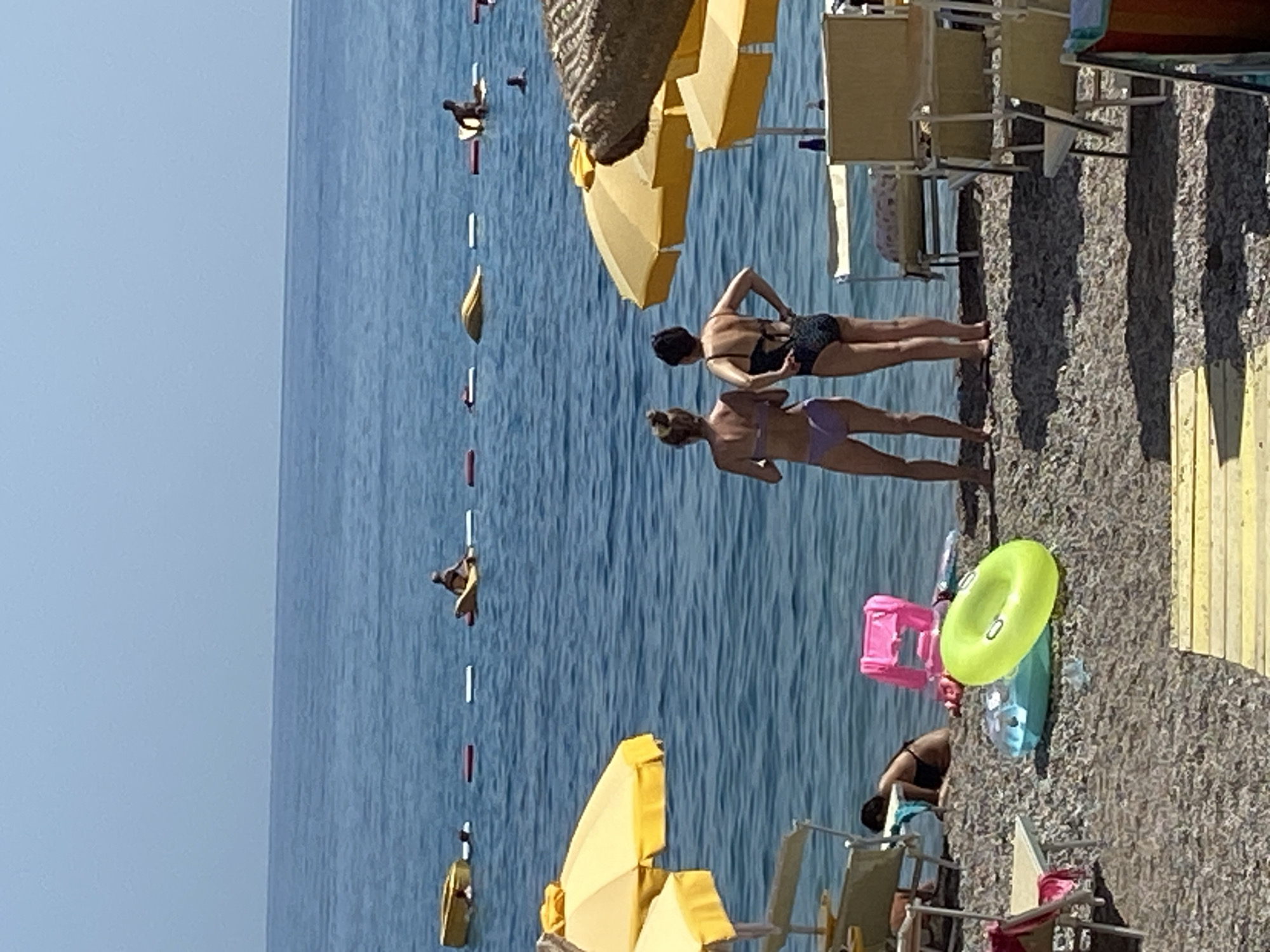 In Prijevor, Montenegro, on a sunny day, a gravel beach meets a calm, blue sea. Two women in bikinis stand facing the water, their backs to the camera. Behind them, yellow umbrellas provide shade over beach chairs. In the sea, several people are visible, some on floating devices, while others swim. In the foreground, inflatable toys, including a bright green ring, lie on the beach, suggesting a day of leisure and recreation.