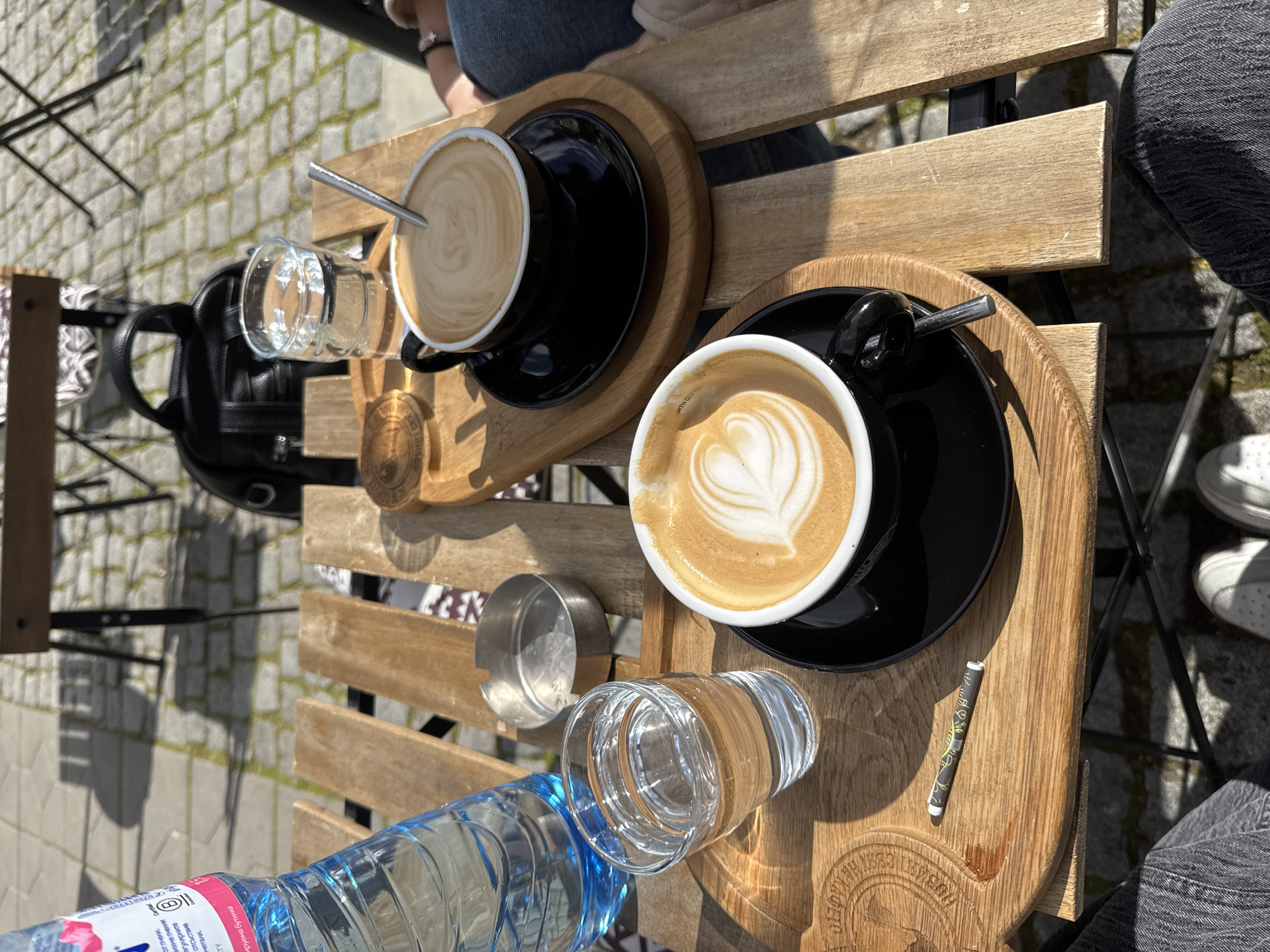 The image shows a table at a café in Goce Delchev, Bulgaria. Two cups of coffee with latte art rest on wooden trays, each accompanied by a glass of water. One cup has a heart design, and the other appears to have a swirling pattern. A blue water bottle is visible on the table, along with a small foil container and a wrapped straw. Part of a black backpack is on one of the seats near the table. Two people are partially visible, one wearing jeans and sneakers, and the other wearing jeans with a light-colored jacket. The table itself is made of wooden slats, and the ground is paved.
