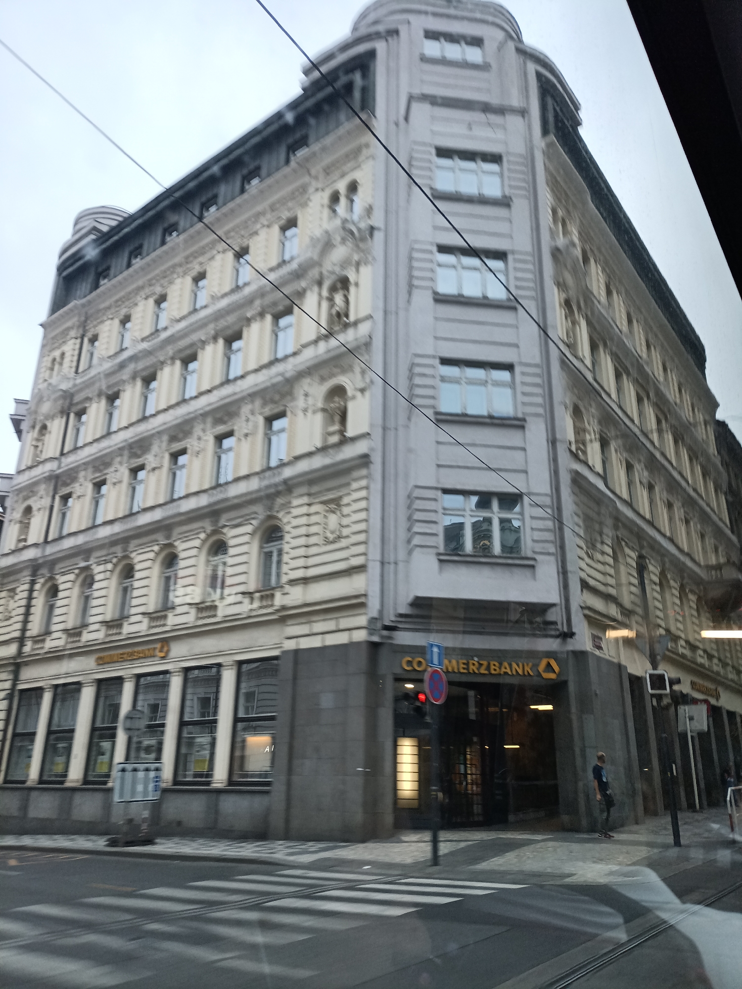 The image captures a street view in Prague, Czechia, featuring the corner of a multi-story building with ornate architectural details. The building displays cream-colored facades with numerous windows, some featuring decorative arched tops. The ground level of the building houses a Commerzbank branch, identifiable by its prominent signage and large glass windows. A pedestrian crossing is visible in the foreground, suggesting a busy urban intersection. Several utility poles and overhead wires are present, indicative of an established city infrastructure. The sky appears overcast, contributing to a muted, cool-toned lighting in the scene. A person is standing near the entrance of the Commerzbank.