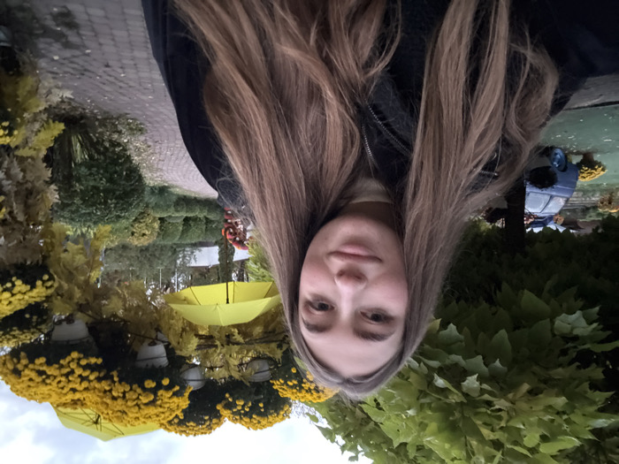 The image displays a young adult female in the foreground, facing the camera with a neutral expression, positioned in an outdoor setting in Lisne, Ukraine. She has long, light brown hair and wears a black jacket over a light-colored turtleneck shirt. In the background, a brick-paved pathway curves to the right. To the left, a large tree with green foliage partially obscures a stationary light blue vintage-style car, which features a roof rack and is surrounded by potted yellow flowers. To the right and center-background, an ornate archway structure is visible, adorned with numerous white pots containing vibrant yellow chrysanthemum flowers and several inverted yellow umbrellas. Manicured green hedges and various shrubs line the pathway, which also shows scattered fallen leaves. The sky is overcast.