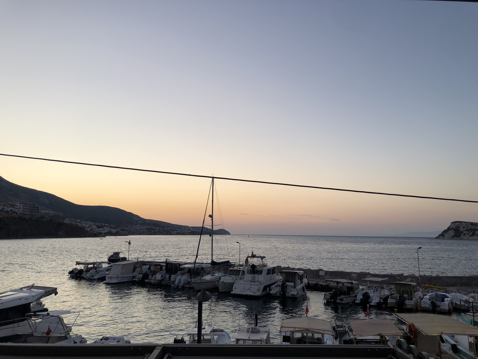 The image shows a harbor in Karaburun, Turkey, at dusk. The sky is a gradient of pale blue to orange, with the sun having recently set. A line of boats of various sizes is moored in the calm water. In the background, a hilly coastline with buildings is visible. The overall scene is peaceful and serene, capturing the beauty of a coastal town at the end of the day.
