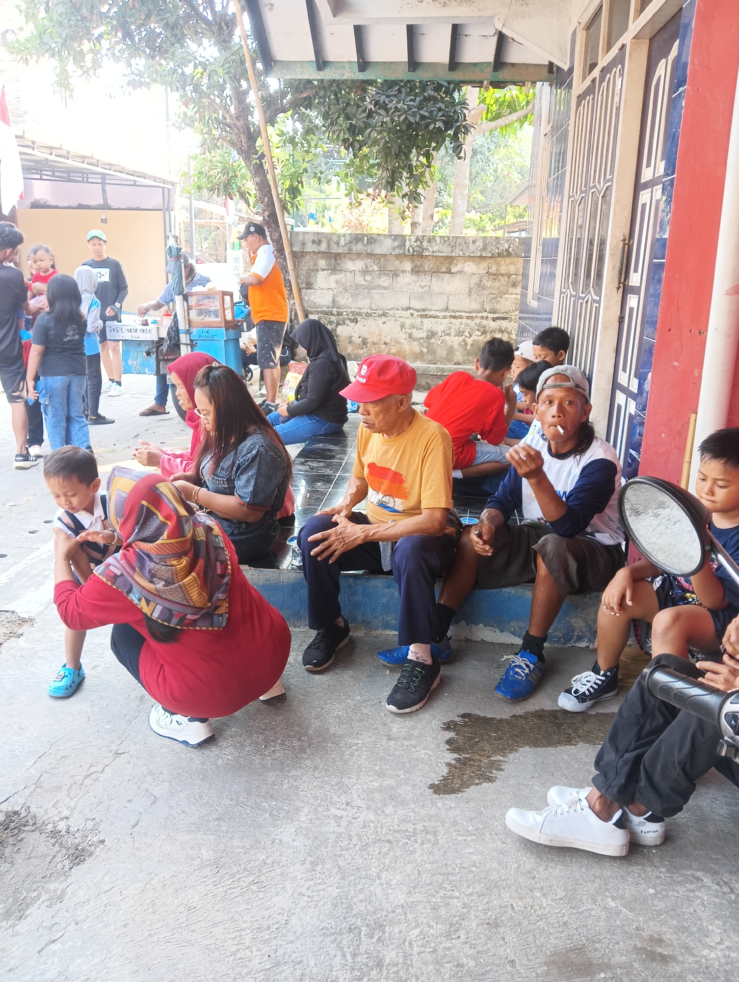 This image depicts a gathering of people, possibly at a street-side food stall, in the city of Mendut, Indonesia. The scene is outdoors, with a building featuring blue and white patterned doors on the right side of the frame, and a concrete wall and stairs in the background. Sunlight casts shadows, indicating it is daytime.
Several individuals are visible, some sitting on steps or the ground, while others are standing. In the foreground, a young child in a red top and a patterned headscarf is interacting with a woman also wearing a red top. A woman in a denim jacket is seated nearby. To the right, an elderly man in a yellow t-shirt and red cap is seated, and next to him, another man in a white and blue t-shirt is smoking a cigarette. A child is seated in front of him, holding a mirror. Several other children and adults are present in the background, with some appearing to be customers or vendors at a stall where "Sosis Okie Okie" is advertised. The overall atmosphere suggests a casual community gathering or a marketplace scene.
