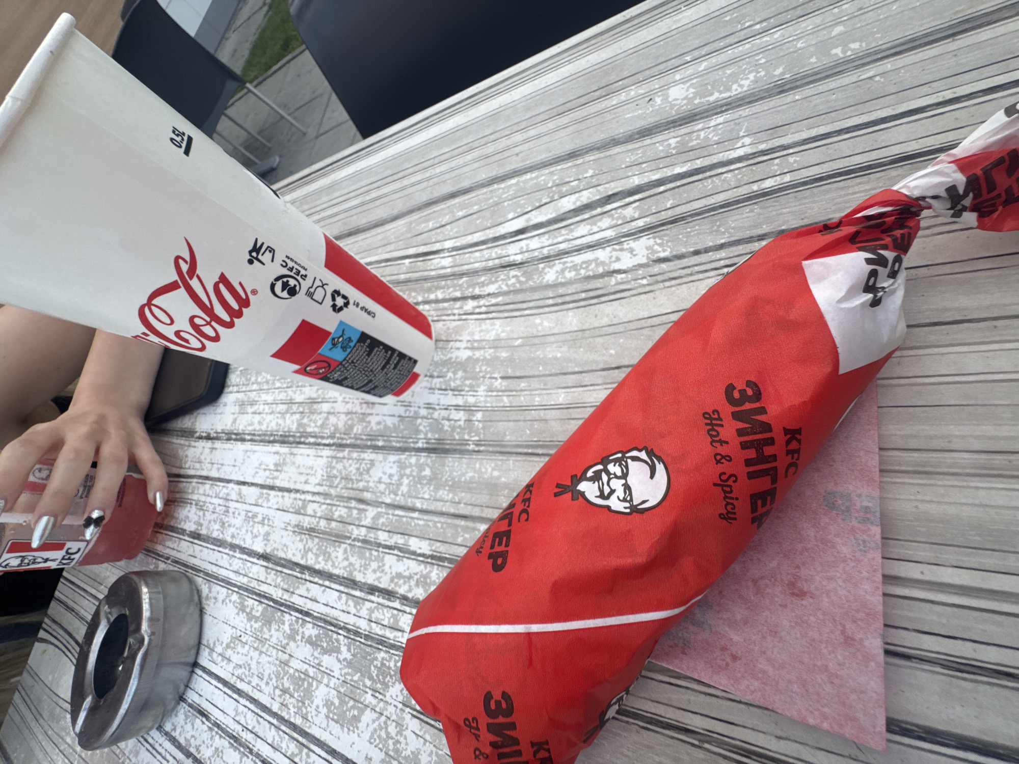 An overhead, slightly angled shot captures a casual dining scene at an outdoor table in Belozem, Bulgaria. The central focus is on two branded beverage cups and a wrapped food item, set against a light-colored table with a distressed, vertically striped surface.
On the left, an oval-shaped, metallic ashtray rests near the top edge of the table. To its right, a clear plastic cup, branded "KFC," contains a reddish-pink liquid with ice. A person's left hand, with metallic silver fingernails (including a black and white floral design on the ring finger), holds this cup. Partially visible beneath the hand is a dark-colored smartphone.
Dominating the right side of the frame is a white paper "Coca-Cola" cup, marked "0.5L" and displaying recycling symbols and a "PEFC" certification. The cup is angled, suggesting motion or dynamic placement.
In the foreground, a red-wrapped KFC Zinger burger, bearing the Colonel Sanders logo and "Hot & Spicy" text, lies horizontally. The wrapping is partially open on the left, revealing a reddish-pink internal paper liner, and twisted shut on the right.
The background is softly blurred, showing a paved ground surface, distant green foliage, and dark chair backs, indicating an outdoor or semi-outdoor dining area under natural daylight.