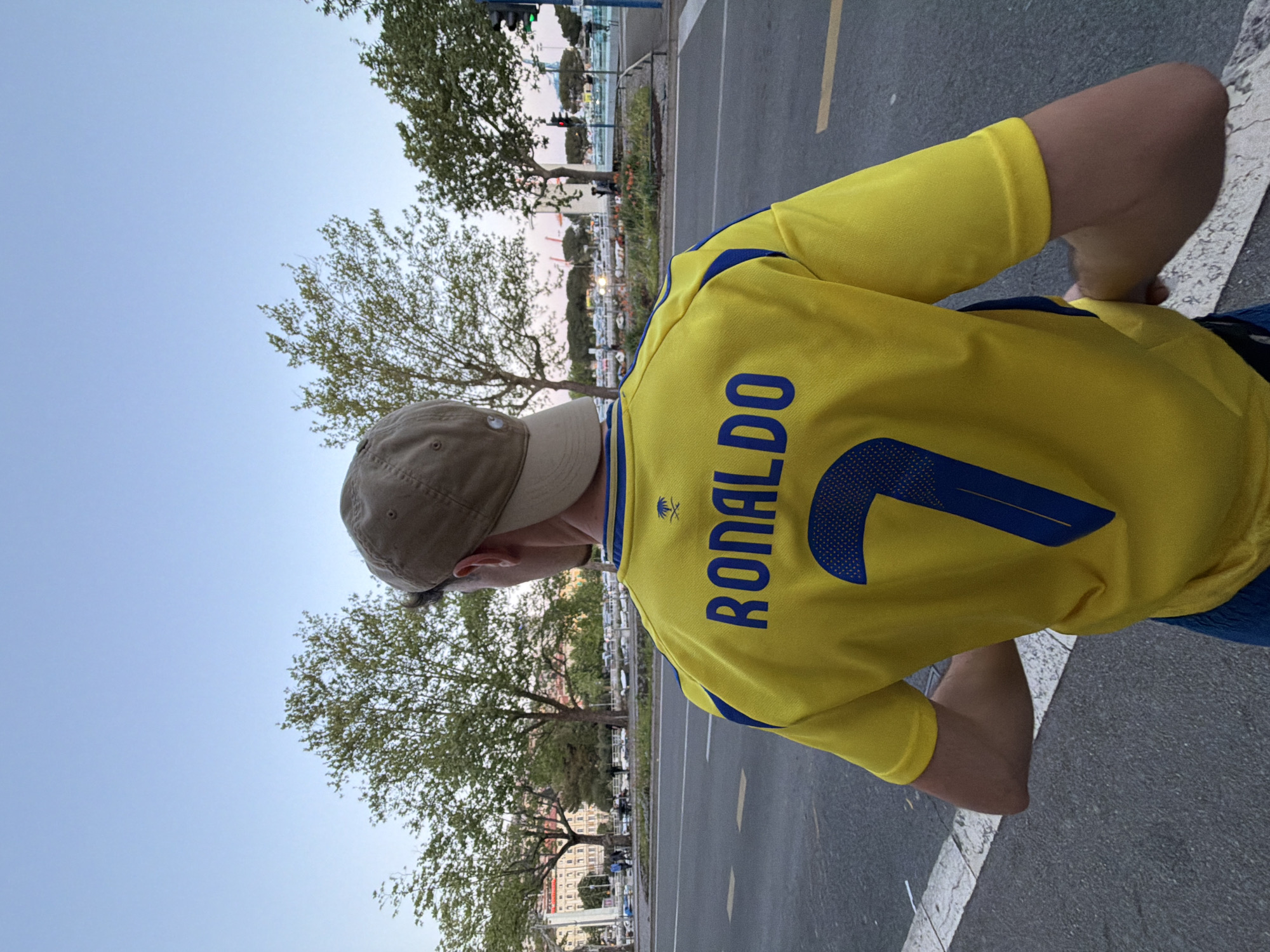 A person wearing a yellow Cristiano Ronaldo soccer jersey and a tan baseball cap is standing on a gray asphalt road. The jersey has the name "RONALDO" and the number "7" in blue lettering on the back. The person is positioned with their back facing the camera, and the left side of their face is partially visible under the cap. The background features trees, buildings, and a marina with boats. A traffic light is visible in the distance. The scene is located in Grad Rijeka, Croatia.