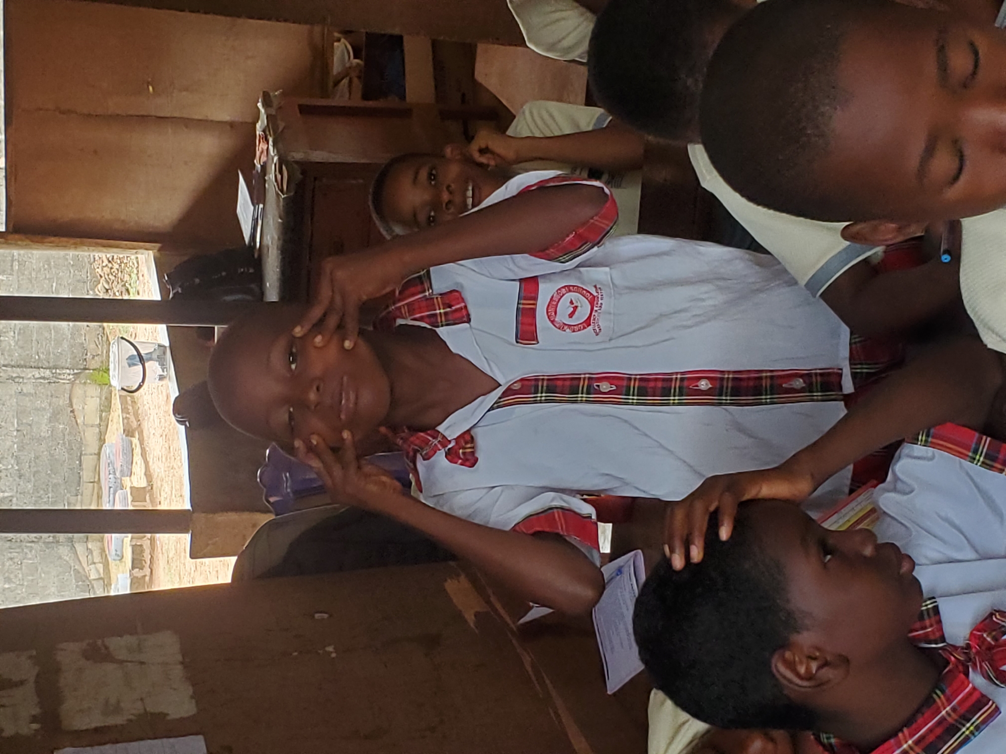A classroom setting in Ukwu-Abwa, Nigeria, depicts a group of students in school uniforms. The uniforms are predominantly white with red and green plaid accents, including a patch on the chest. One student is actively engaging with the camera, holding their hands to their face. Other students are seated, some with their heads resting on their hands. The room has a rustic appearance with visible exterior elements like windows, showing parts of the surrounding area. The scene appears to be taken in the afternoon or early morning, with natural light illuminating the classroom.