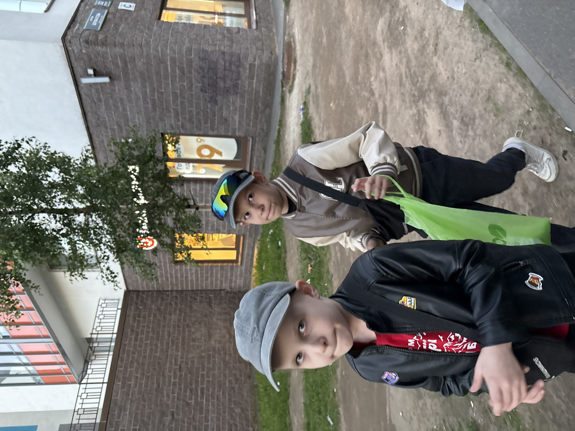 Two young boys are walking outdoors in a city environment. The boy in the foreground, on the left, is wearing a gray baseball cap, a black leather jacket over a red t-shirt, and has his hands clasped in front of him. He is looking towards the camera with a neutral expression. The boy behind him, on the right, is wearing a gray baseball cap with reflective sunglasses perched on top, a brown and cream-colored bomber jacket, and dark pants. He is carrying a bright green plastic bag in his right hand and looking to his left. They are walking on a paved surface next to a grassy area. In the background, there is a multi-story building with brick and white facades, featuring red and white striped architectural elements on an upper floor. A tree with green foliage is situated in front of the building. A street sign with Russian text and numbers "22" and "C" is visible on the building's corner. The lighting suggests it is daytime, possibly late afternoon or early evening.