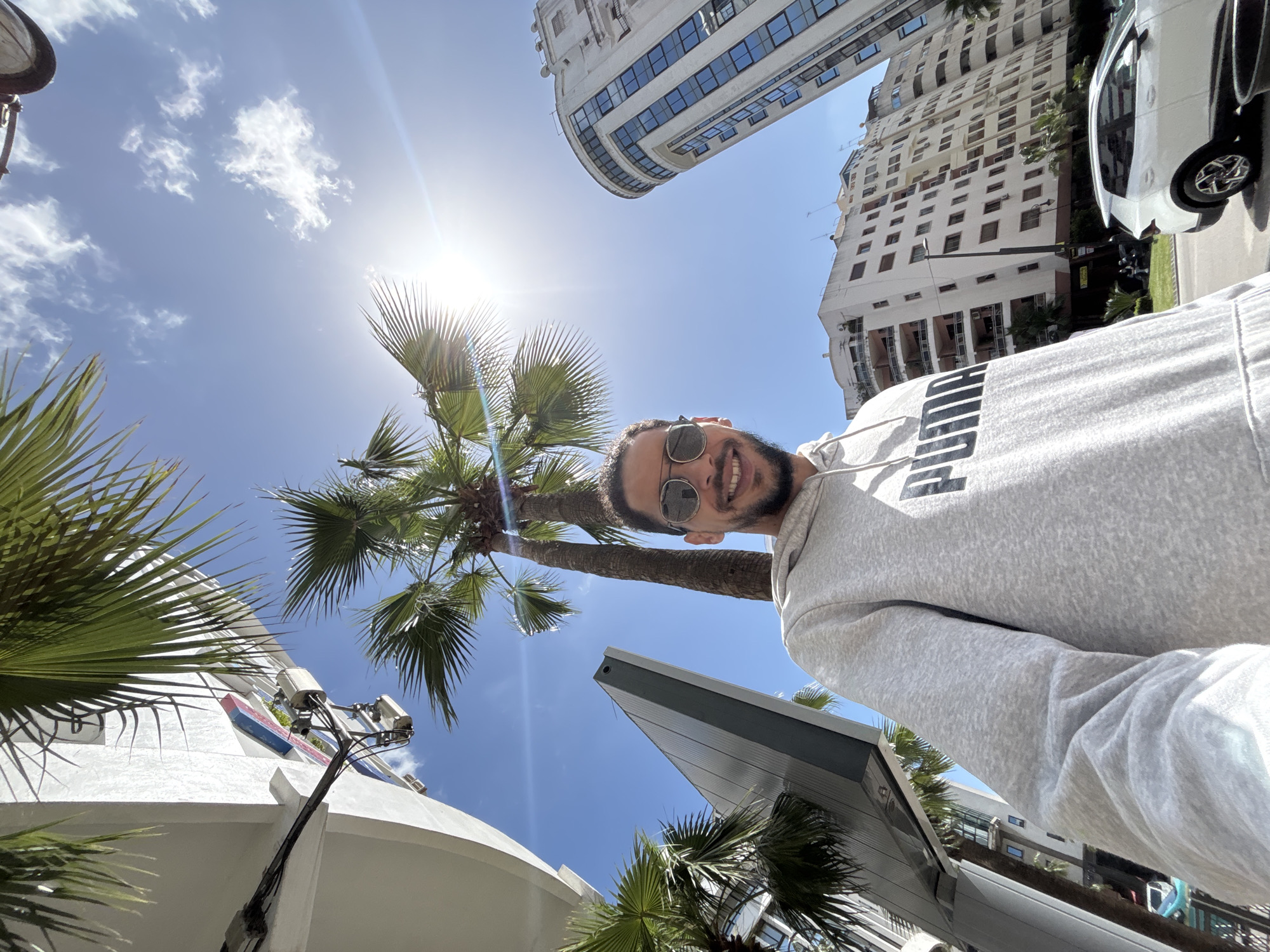 The image depicts a male individual captured in a low-angle selfie format, smiling and wearing reflective sunglasses and a grey "PUMA" brand hoodie. The scene is set outdoors under a bright blue sky with scattered white clouds, featuring a prominent sun glare in the upper right quadrant.
The environment is an urban setting in Tanger, Morocco. Multiple tall palm trees with green fronds are visible, framing the sky and partially obscuring architectural elements. On the left, a modern, white building with mounted technical equipment and cables is partially visible, alongside a distinctive grey, angular architectural structure. On the right, a prominent, multi-story building with a rounded facade and numerous blue-tinted windows rises upwards. Further back, a large, multi-story white residential building with balconies is present. A white SUV is visible on a street or roadside in the lower right background, behind the individual, next to a landscaped area featuring grass and additional trees. The overall impression is a sunny day in a contemporary city district.