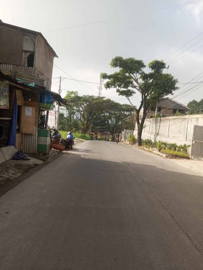 A quiet, paved road winds through a residential area. A small shop with a corrugated metal awning sits on the left, with a motorcycle parked nearby. Across the road is a tall concrete wall lined with green plants. Trees canopy the road further down, creating a tunnel-like effect. Several people on motorbikes are seen in the distance. Power lines stretch across the sky. A tall communications tower is visible in the background. The sky is overcast.