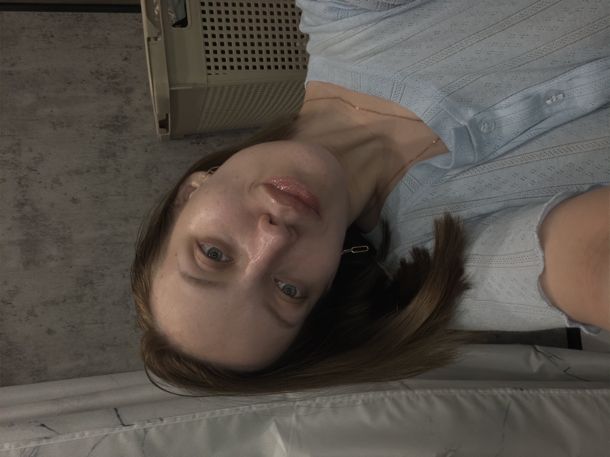 A woman with fair skin, blue eyes, and light brown hair is pictured in a selfie. She is wearing a light blue, button-down shirt and a gold necklace. The background includes a gray wall, a white object resembling fabric or a sheet, and a beige plastic storage container. The location is not possible to determine from the image.
