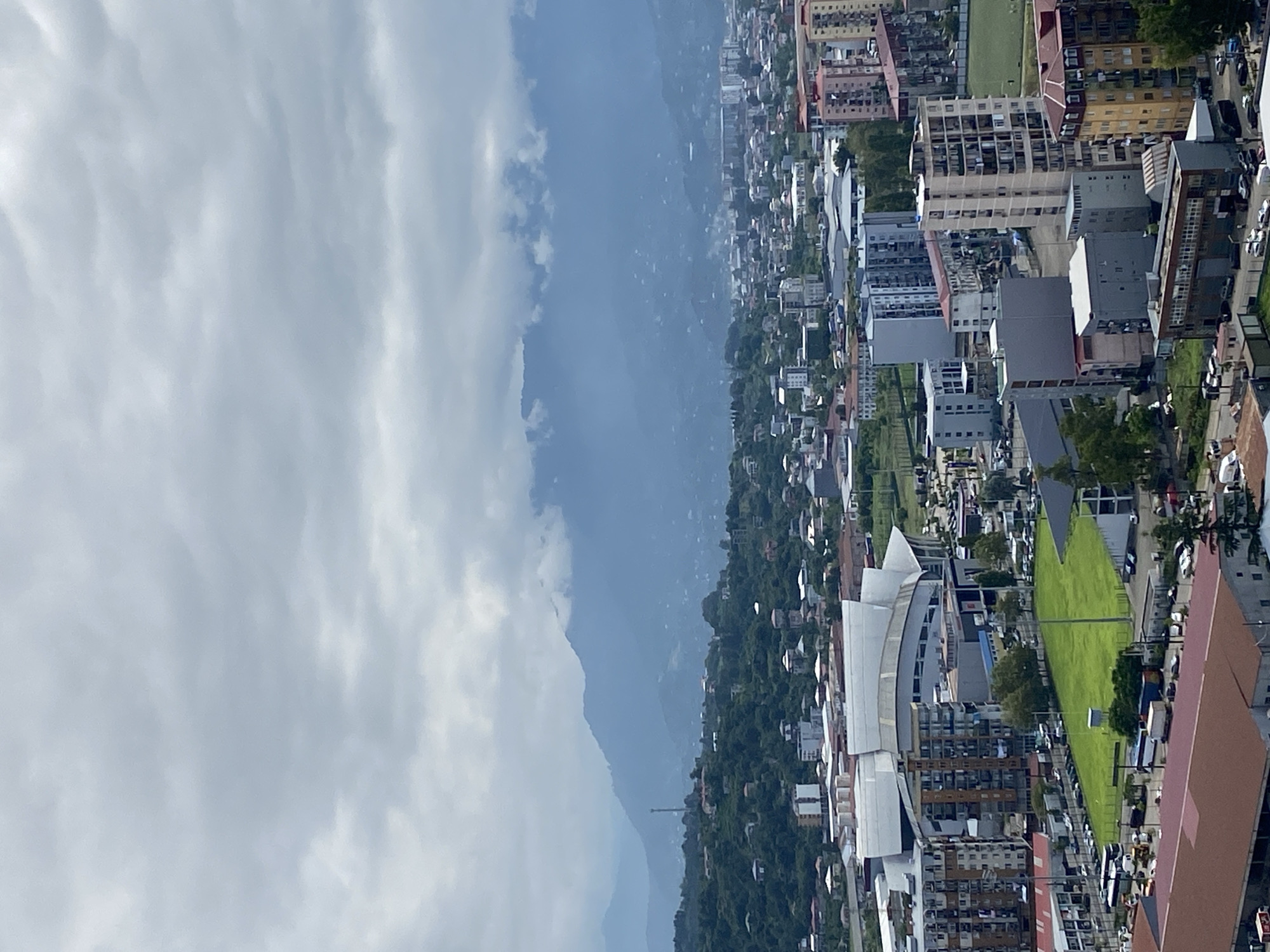 An aerial, wide-angle view captures a cityscape with mountains in the background under a cloudy sky. In the foreground, a large stadium-like structure with a white, curved roof dominates the scene, bordered by several multistory buildings and a green, rectangular field. Numerous cars and buses are visible in parking areas and on roads. The middle ground shows a dense urban area with various apartment buildings of different heights and architectural styles. Further back, the city extends towards the base of lush green hills, dotted with smaller buildings and a telecommunications tower. The farthest background is characterized by layered, mist-covered mountains, their peaks obscured by thick clouds. The overall impression is of a developing urban center nestled against a natural mountainous backdrop.