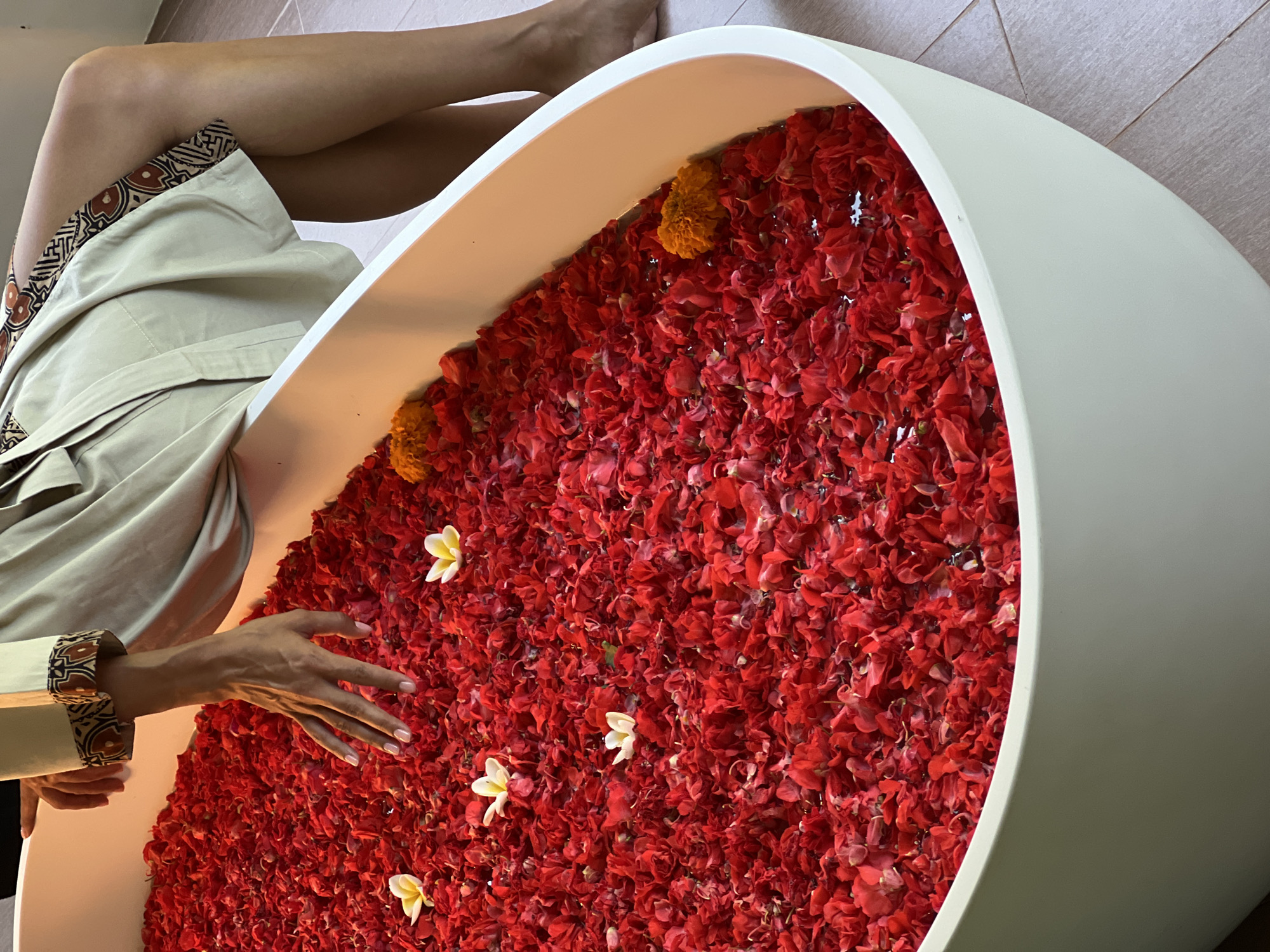 A close-up, high-angle shot shows a white bathtub filled with red rose petals and a few white and orange flowers. A person's hand, with manicured nails, reaches into the petals. The person is wearing a light-colored robe with decorative trim on the sleeves. The scene appears to be in a bathroom, possibly in a hotel or spa, in the Ubud District of Indonesia, a region known for its cultural and natural beauty. The floor tiles are visible in the lower right corner of the image, suggesting an indoor setting.