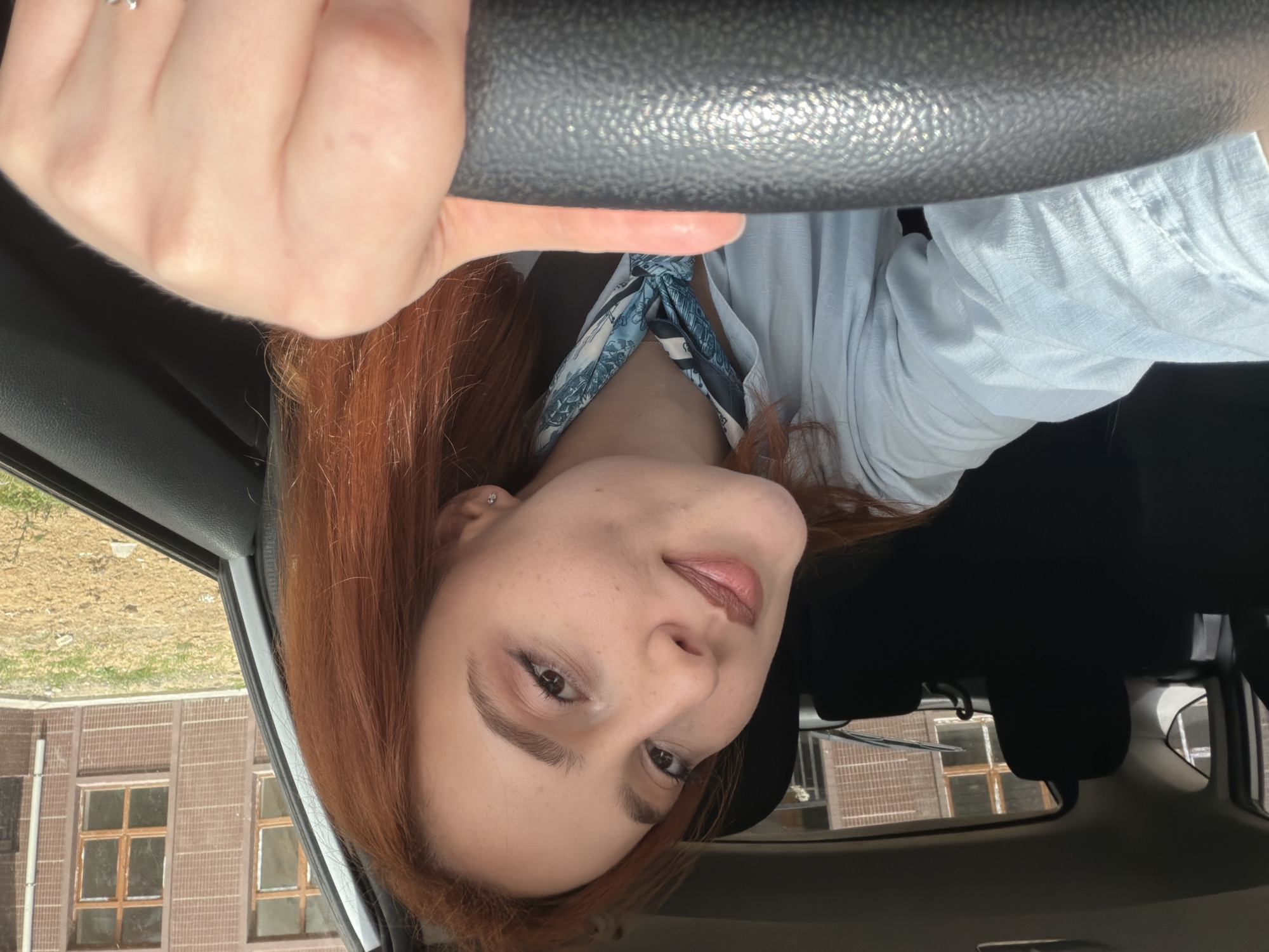 A woman with red hair is sitting in the driver's seat of a car, holding the steering wheel. She is wearing a light blue shirt and a patterned scarf. The exterior view through the car windows shows a building with windows and an adjacent patch of land, suggesting a location in an urban area. The building architecture appears to be reminiscent of the architecture in Tbilisi, Georgia. The image is taken at an unusual angle, perhaps from below.
